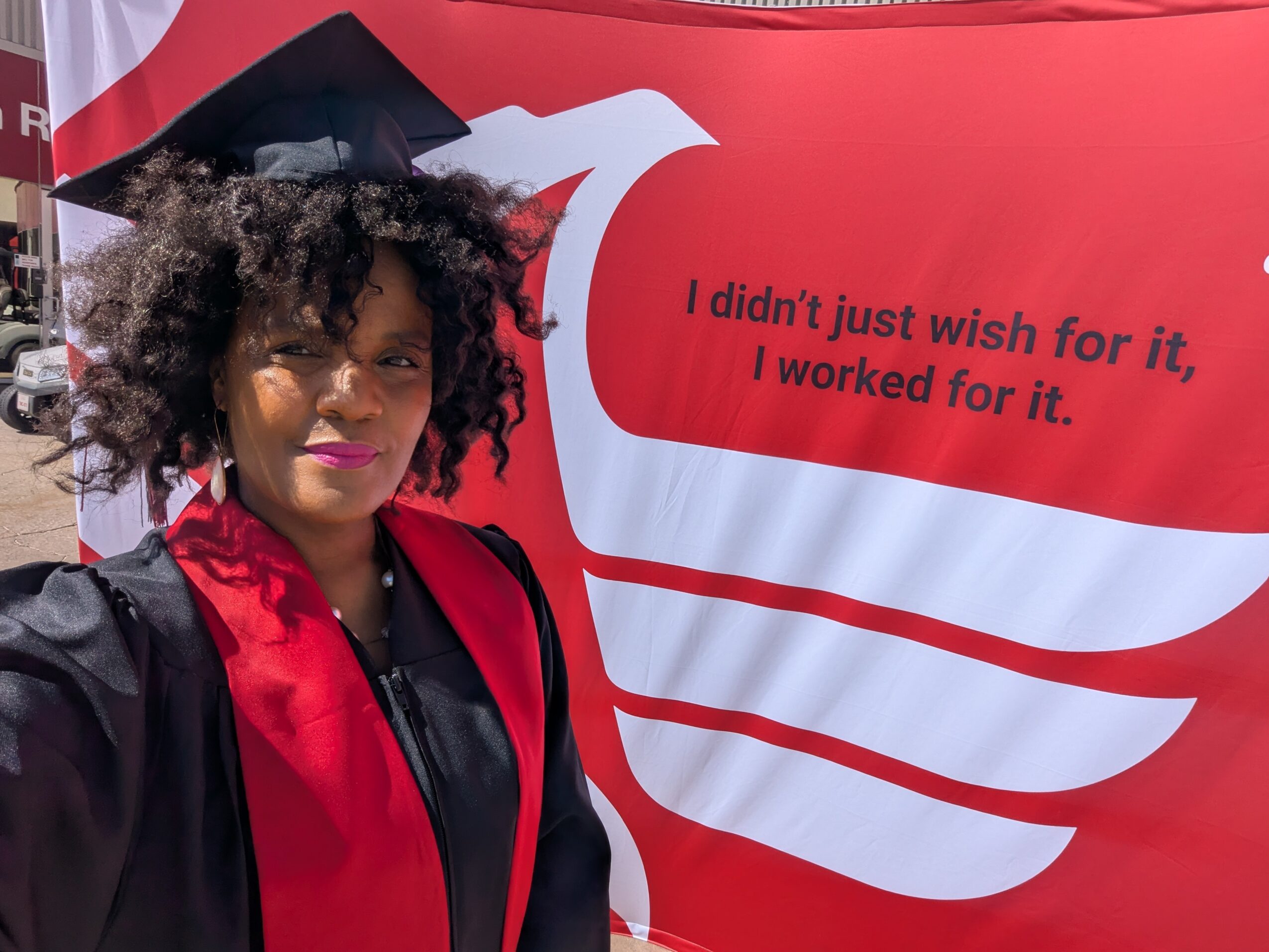 Woman in graduation gown and cap taking selfie in front of a red banner with white text.