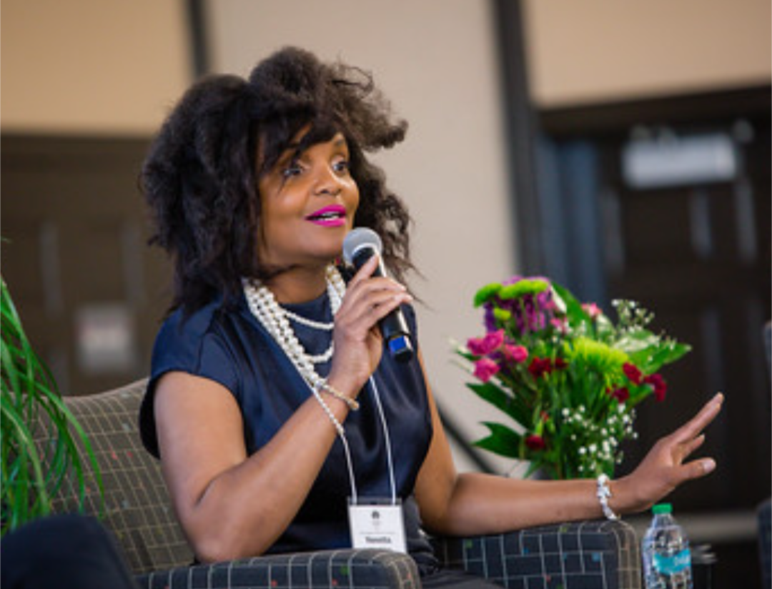 Woman speaking into microphone with flowers on table, wearing dark dress and pearl necklace.