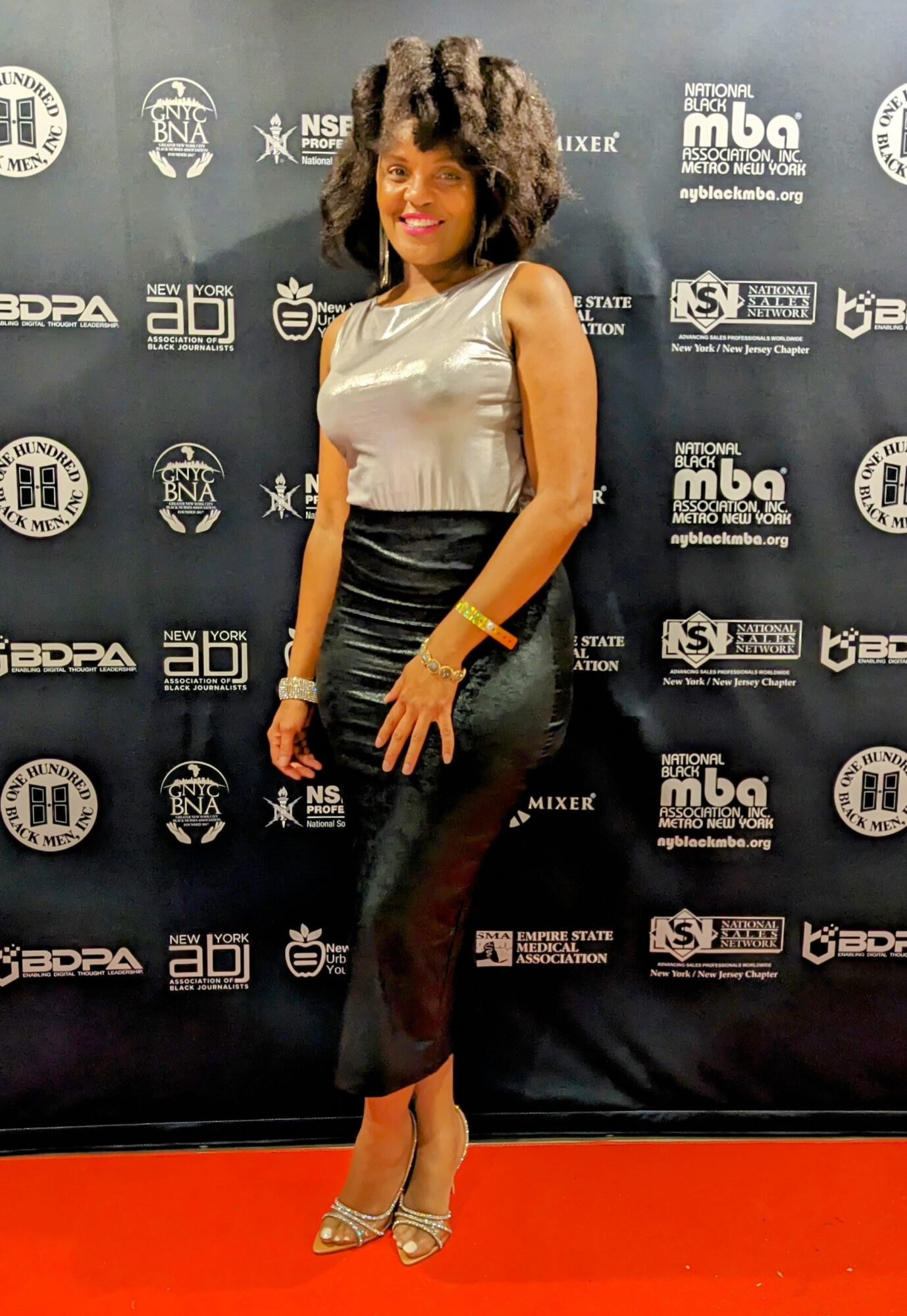 Woman in silver top and black pants standing on red carpet in front of a black backdrop with logos.