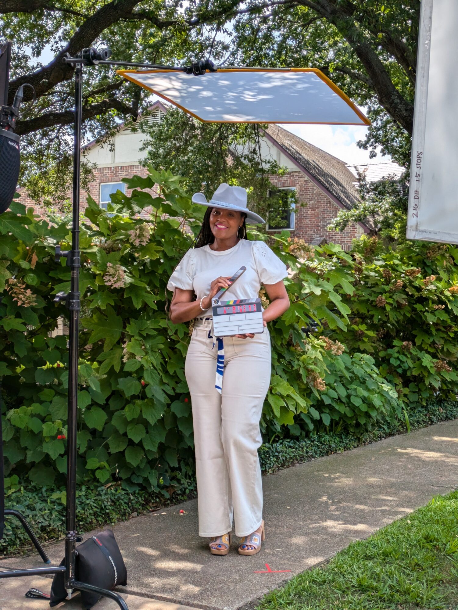 Woman in white hat and outfit holding a book outdoors near green bushes and a brick building.