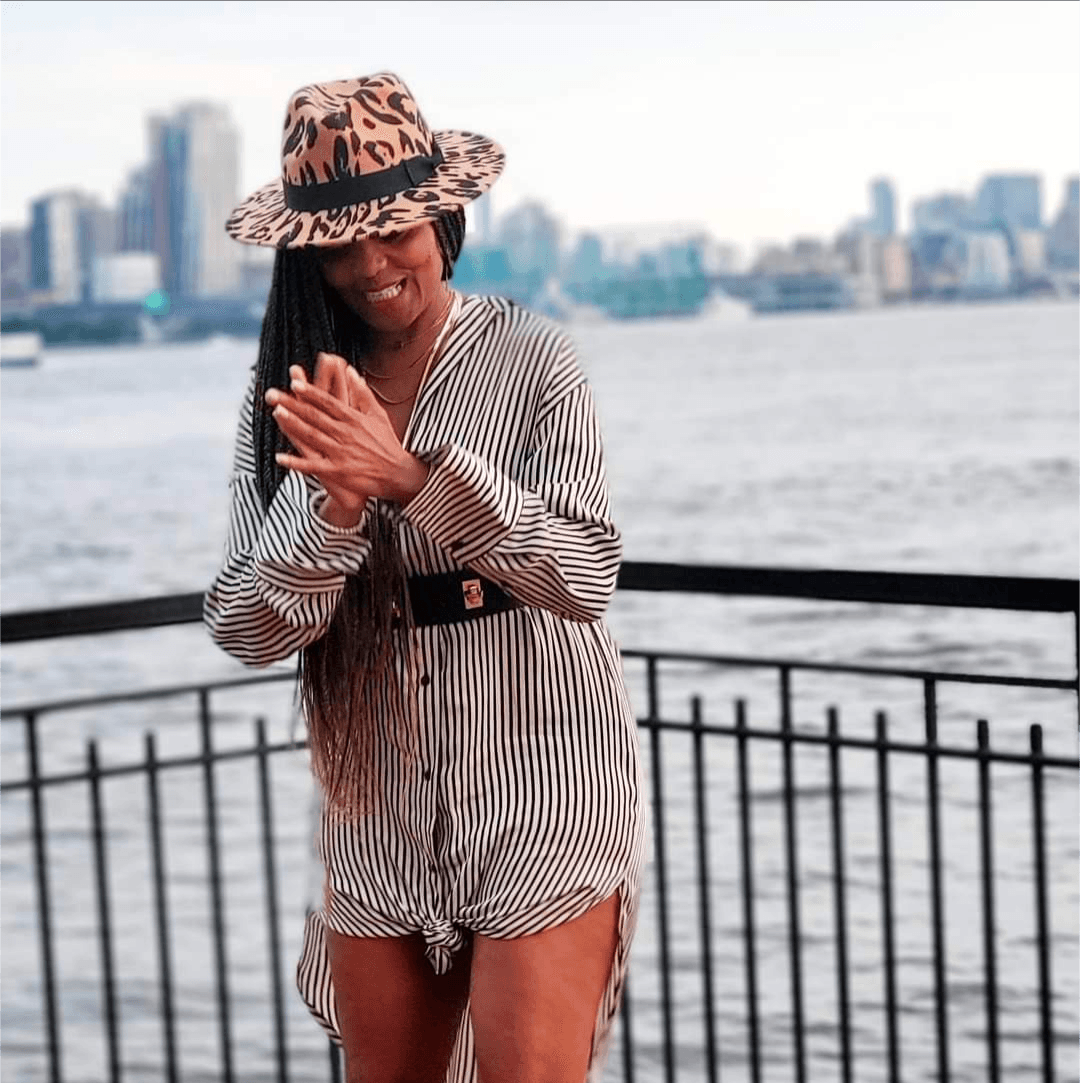 Woman wearing a striped outfit and leopard print hat, smiling with hands clasped, standing by water with city skyline.