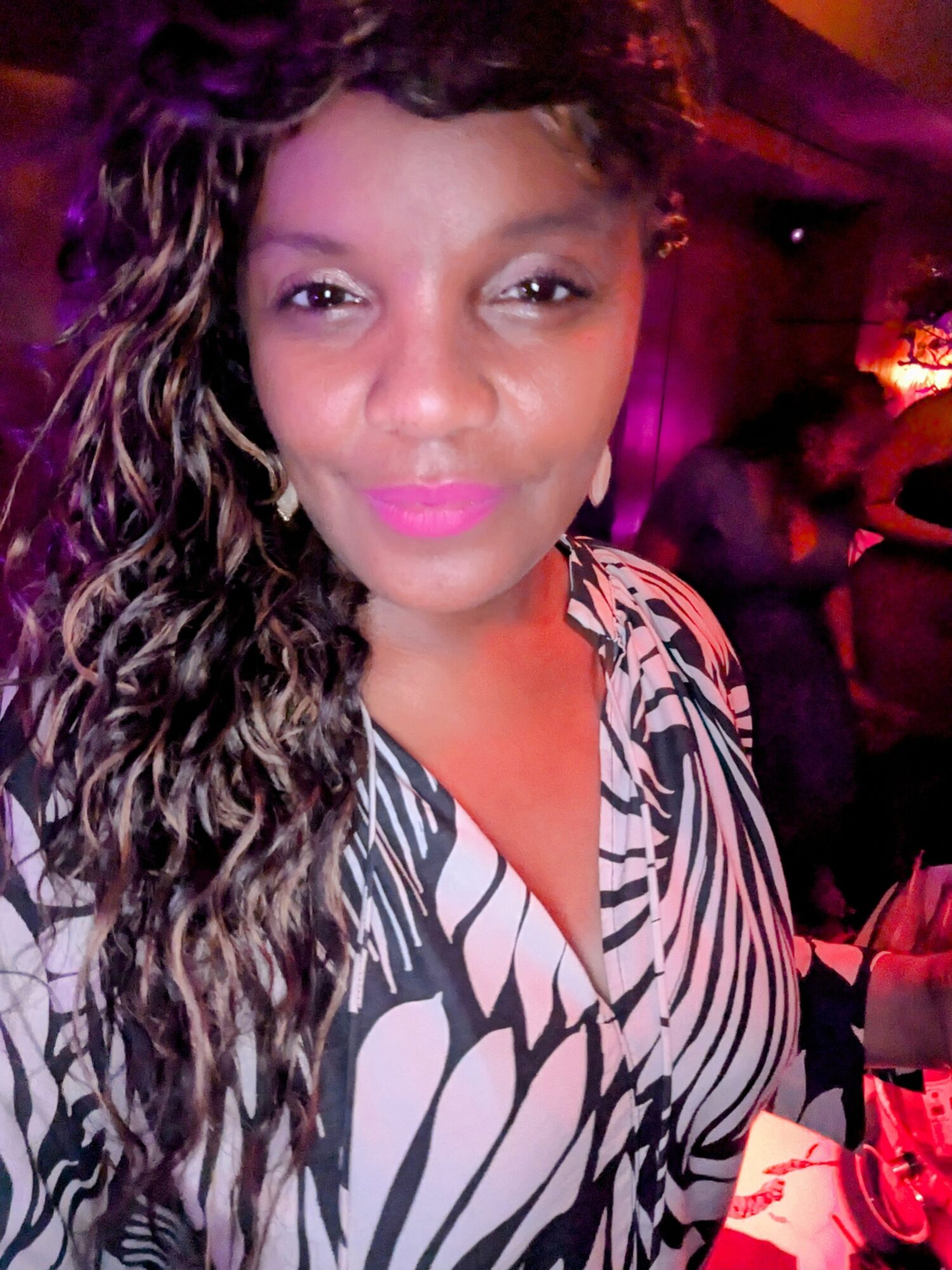 Woman with long, curly hair and pink lipstick in a patterned top at a dimly lit event.