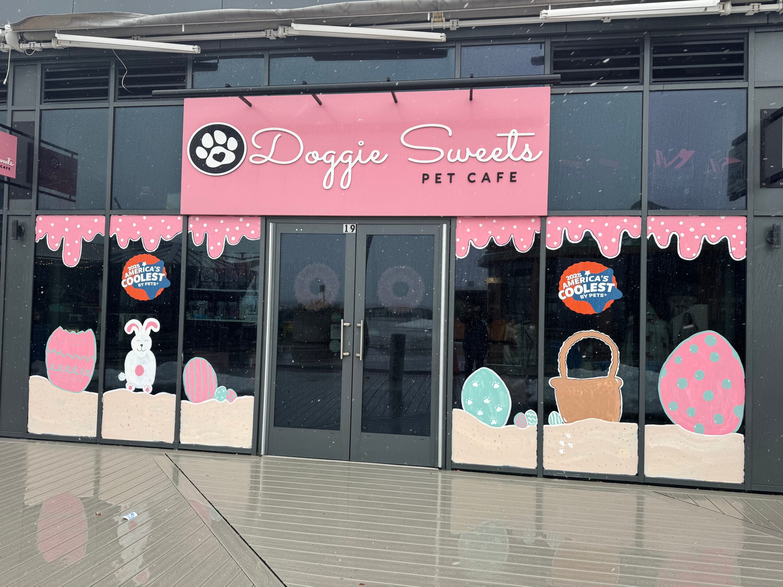 Storefront with pink sign reading 'Doggie Sweets Pet Cafe' and window decorations of a dog, eggs, basket, and pink patterns.