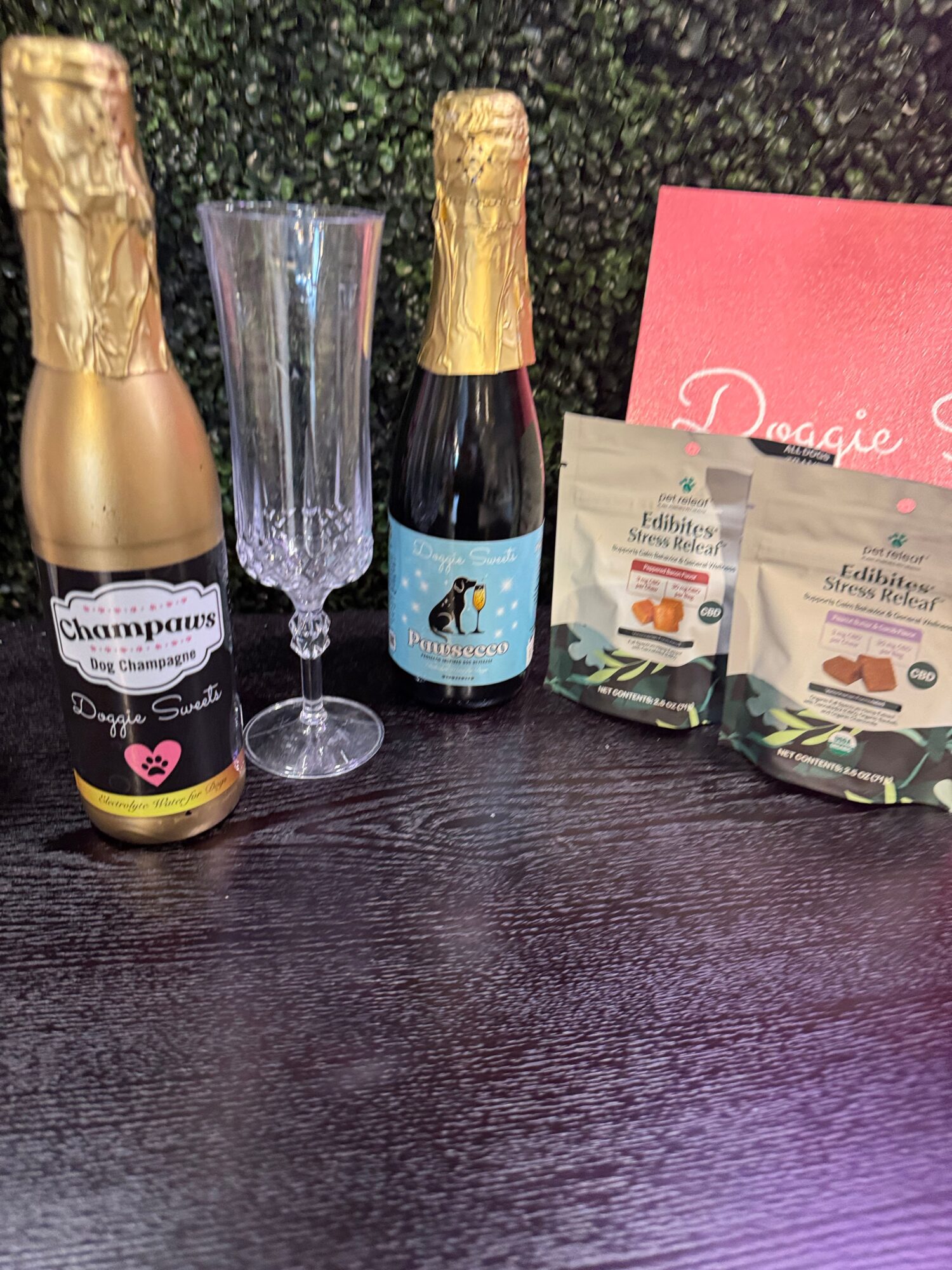 Two bottles of champagne, a tall empty glass, and two packets of snacks on a dark surface with a pink card in the background.