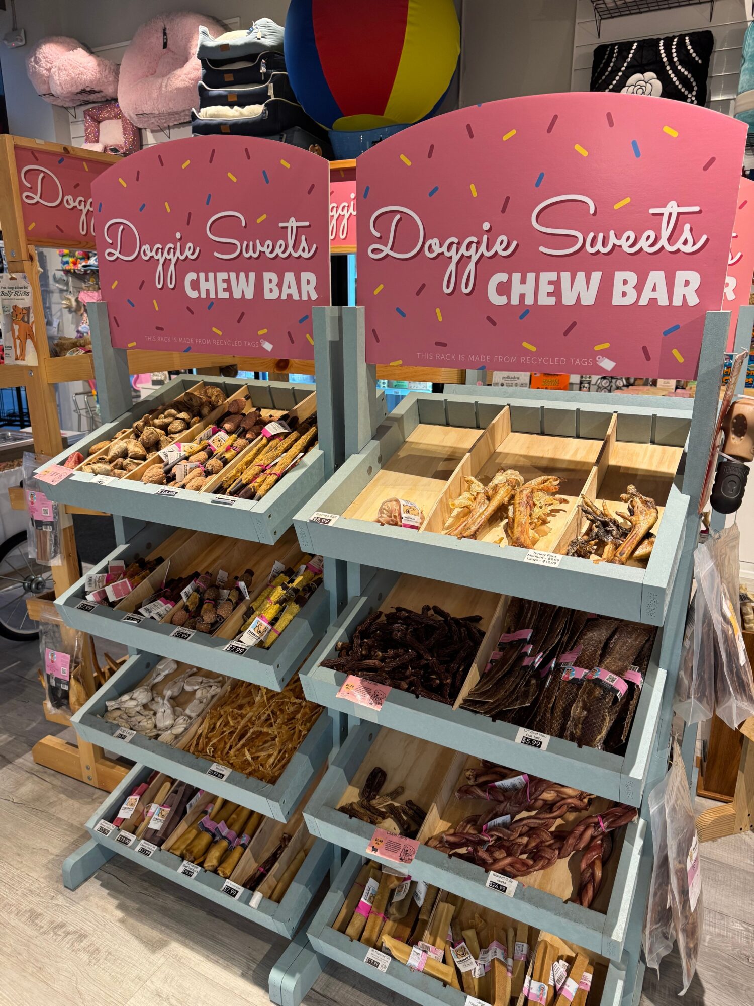 Display shelves with dog treats and chews, pink signs reading 'Doggie Sweets Chew Bar', in a store.