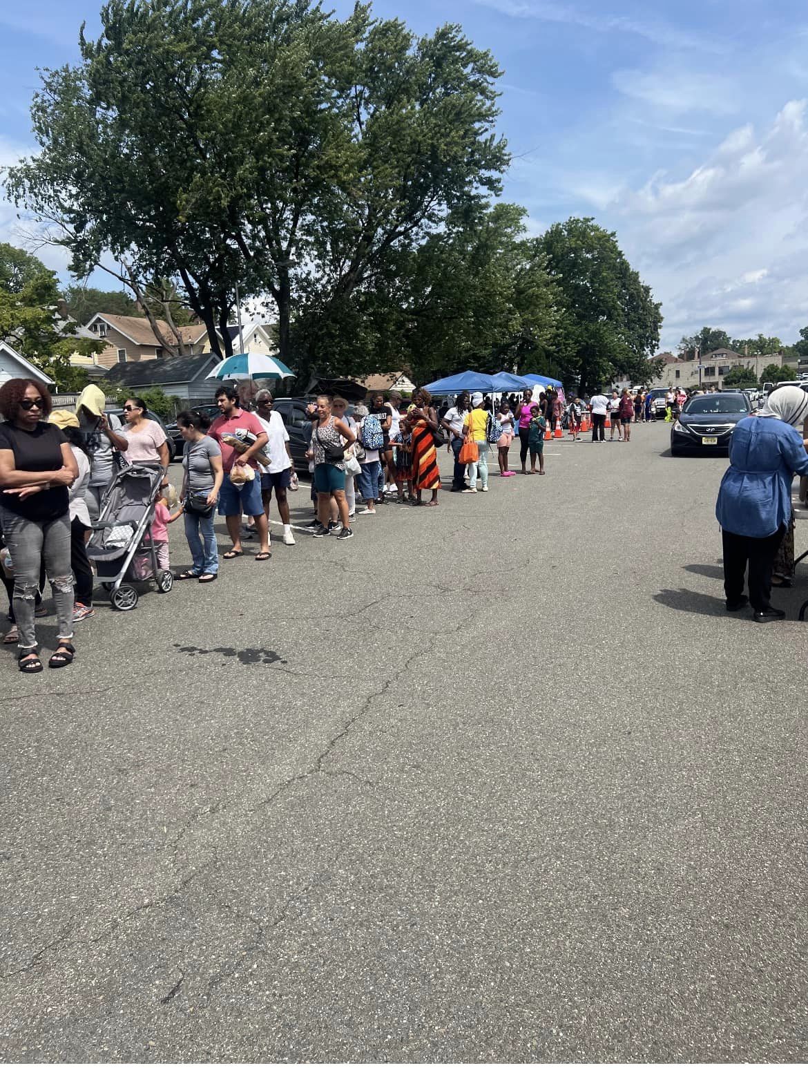 People standing in a long line outdoors on a sunny day, with trees and tents in the background.