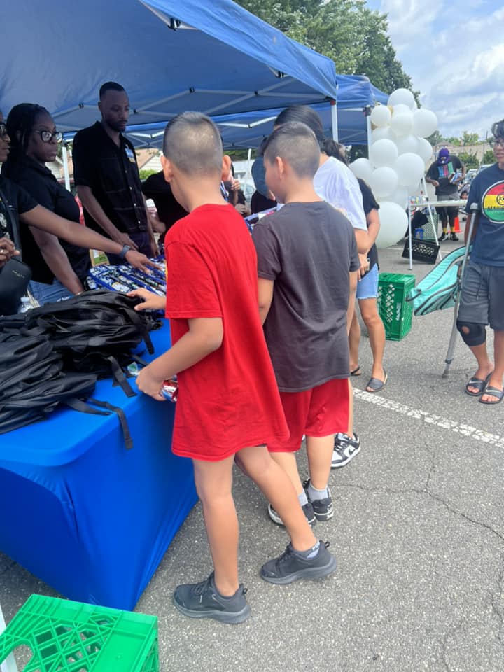 Children and adults gather around a table with black items under a blue canopy outdoors.