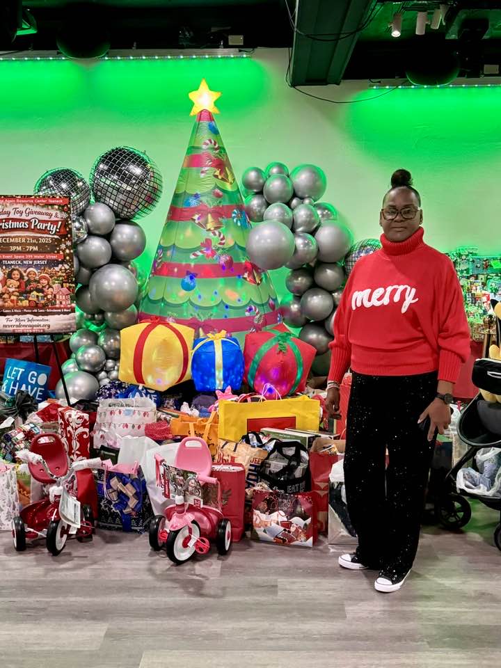 Woman in red sweater standing next to Christmas decorations and presents, decorated Christmas tree with ornaments and balloons in background.