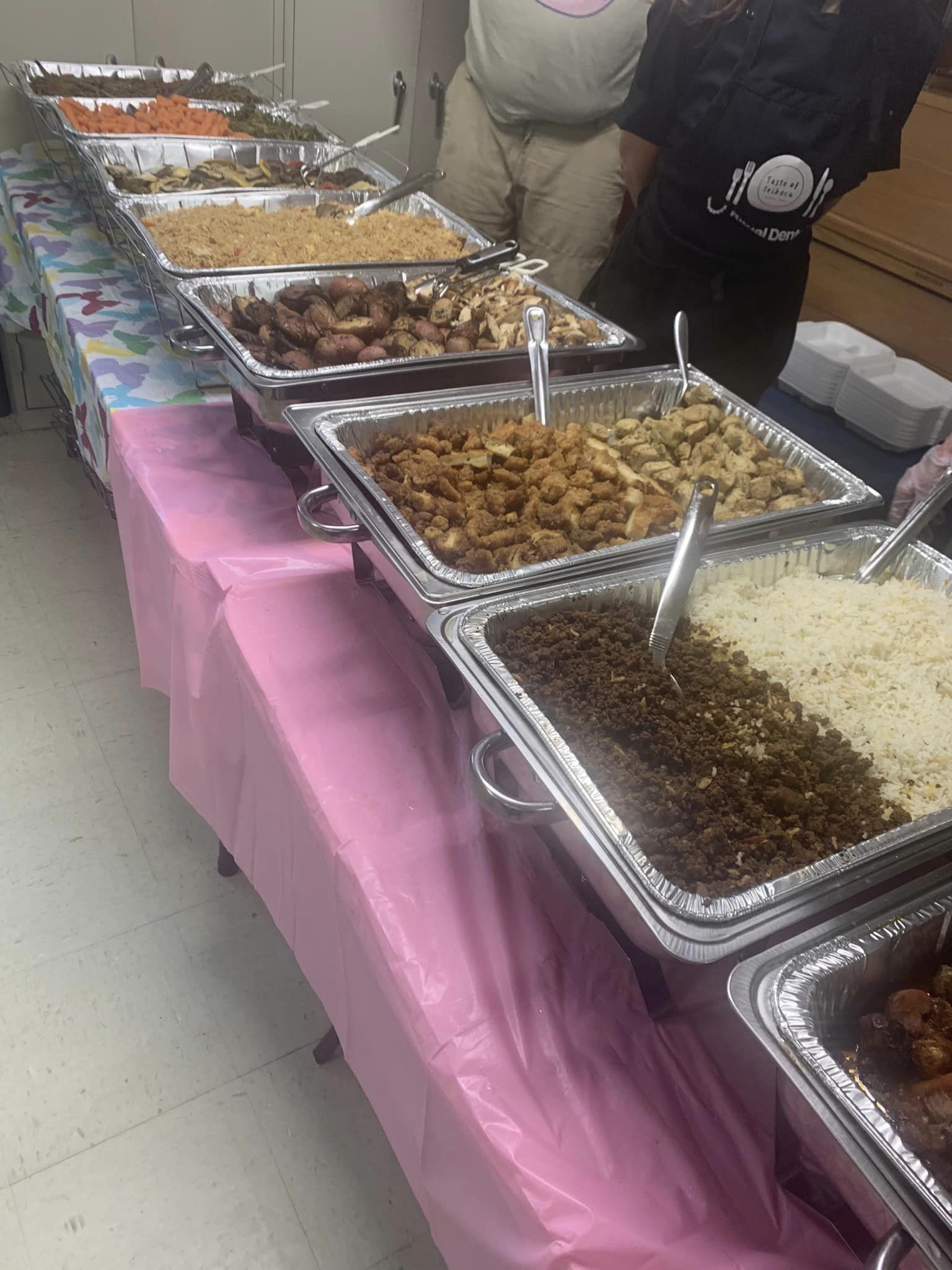Buffet table with various dishes in metal trays, pink tablecloth, people in background, food includes rice, beans, meat, and vegetables.