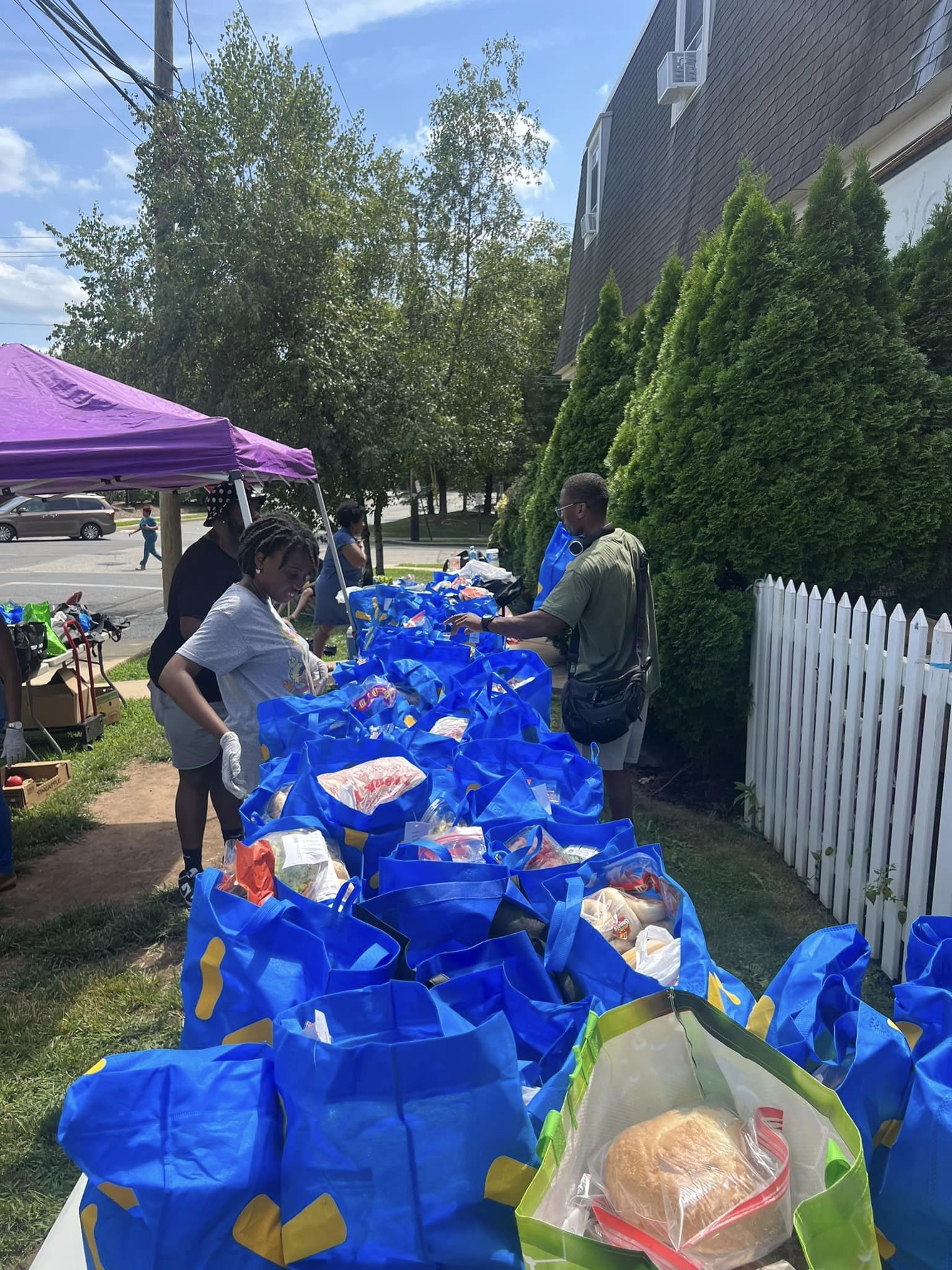 People gather around a table with blue bags and food items outdoors, with trees, a purple canopy, and a white fence nearby.