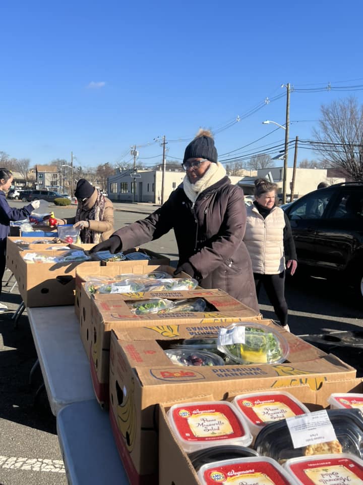 People shopping at an outdoor market with boxes of food on a table, in a sunny urban setting.