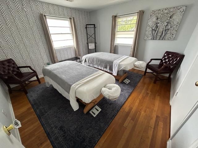 Two beds in a bedroom with two windows, chairs, a painting, and a dark rug on wooden floor.