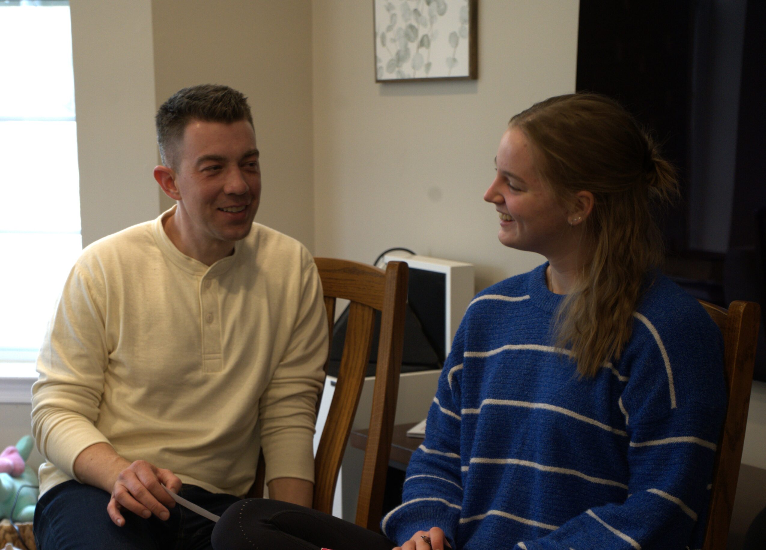 Man and woman sitting and talking indoors, smiling at each other, wooden chairs, framed picture on wall.