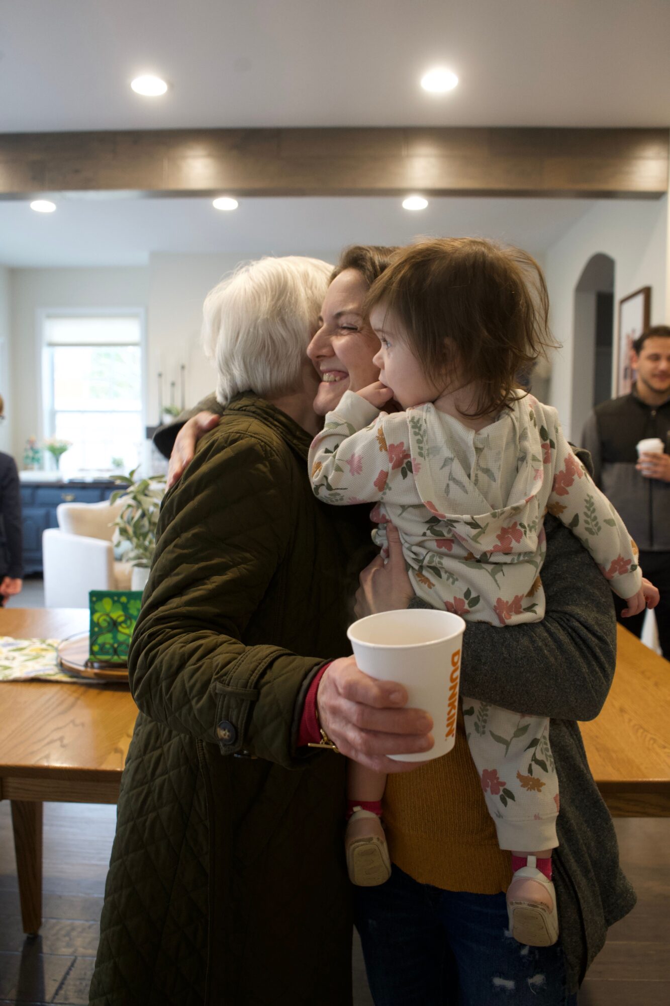 Three people hugging in a room with a wooden table and a window in the background.
