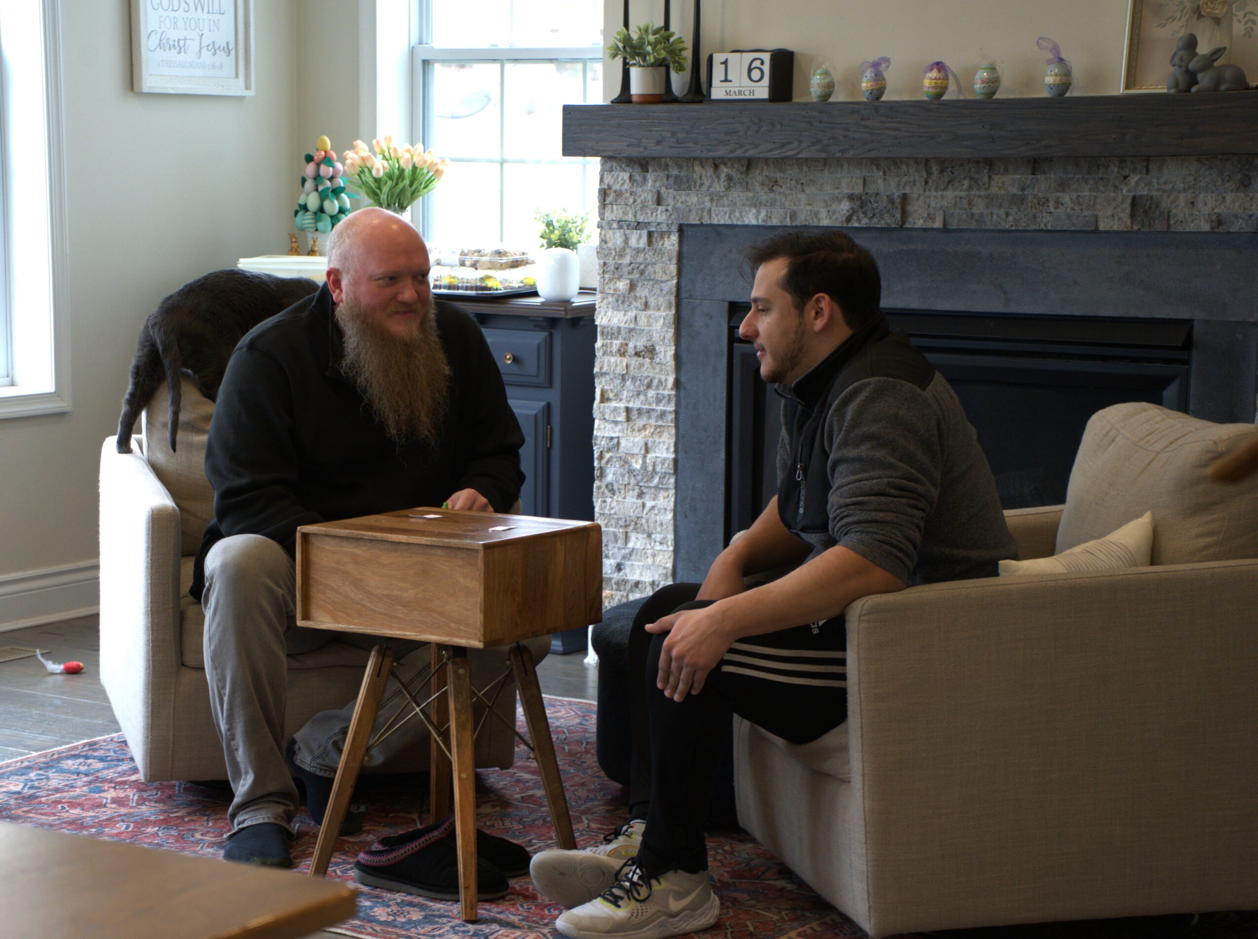 Two men sit and converse in a living room with a fireplace, sofa, and window with flowers on the windowsill.