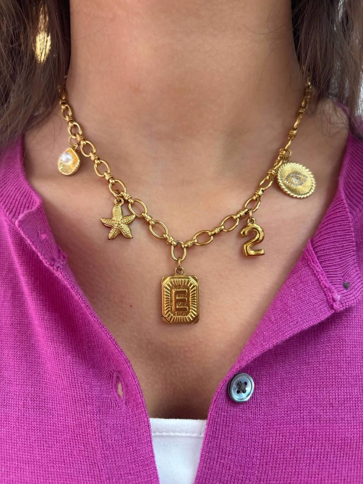 Person wearing a gold chain necklace with various charms, pink top, and white shirt underneath.