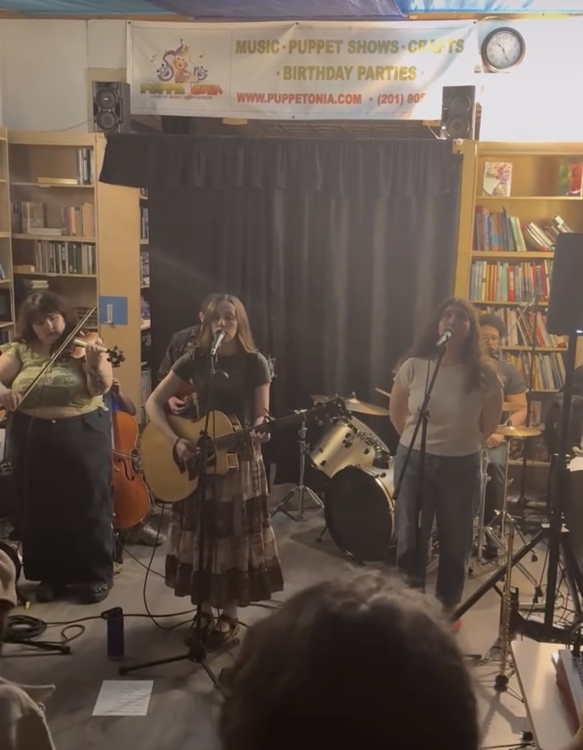 Three women perform on stage with musical instruments in a library setting, bookshelves visible on sides.