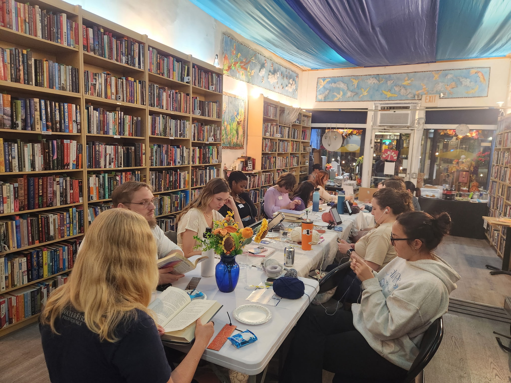 People sitting at a long table in a bookstore, reading and talking, with bookshelves and colorful decorations in the background.