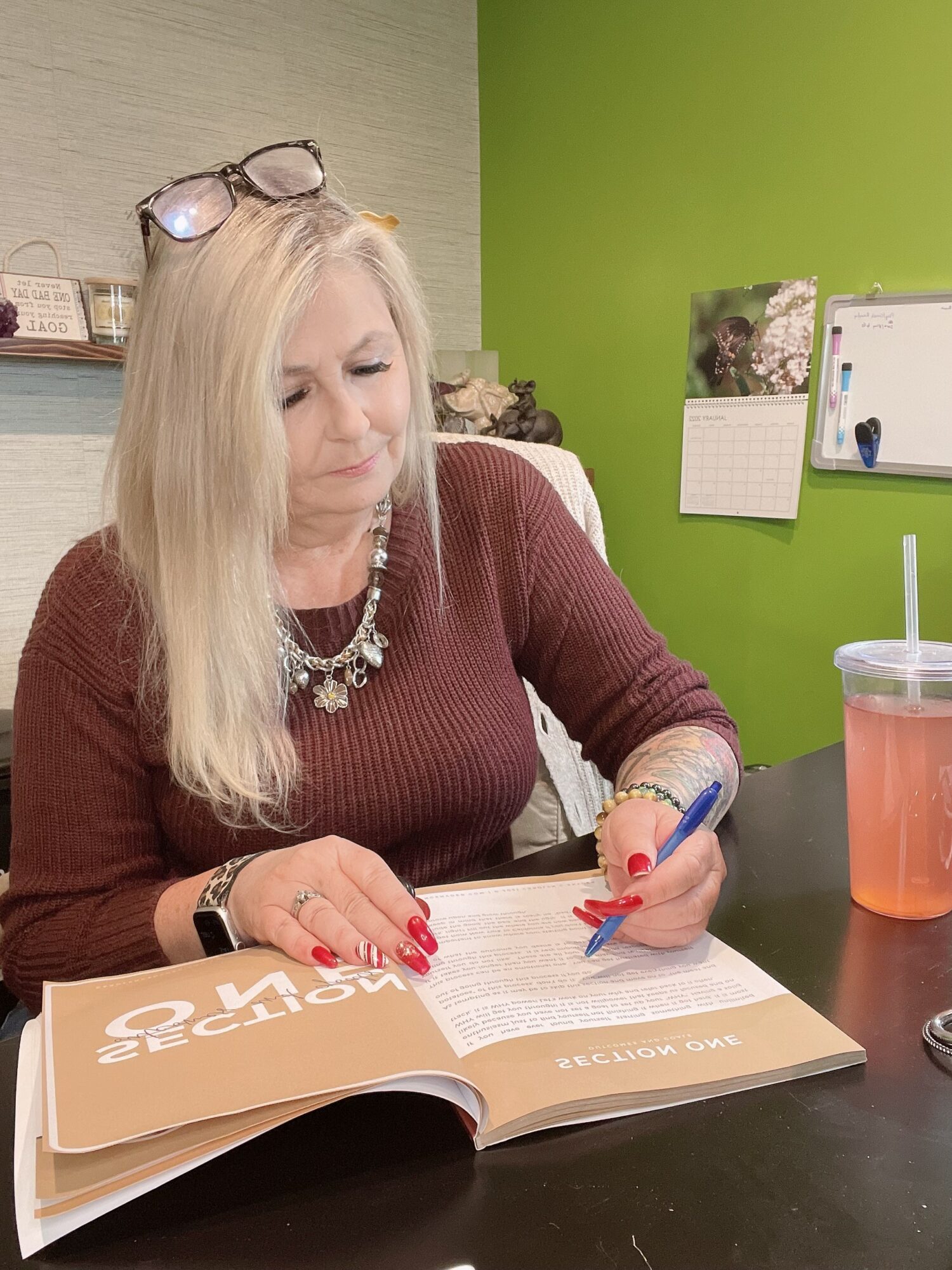 Woman with blonde hair reading a magazine at a desk, with a green wall and calendar in background.