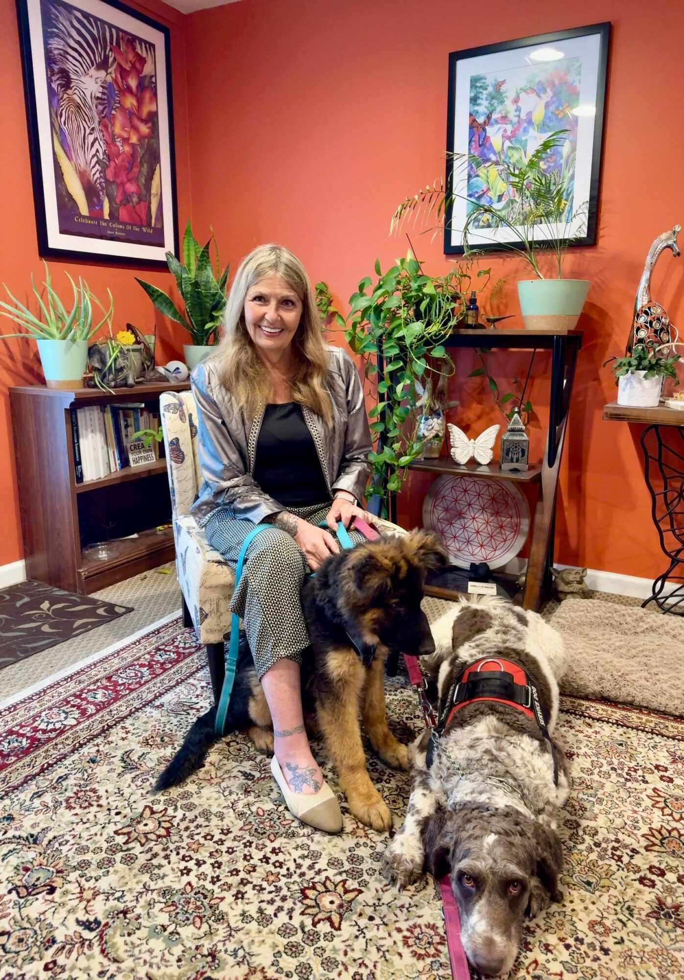 Woman sitting on a chair with two dogs in a room decorated with plants and framed artwork on orange walls.