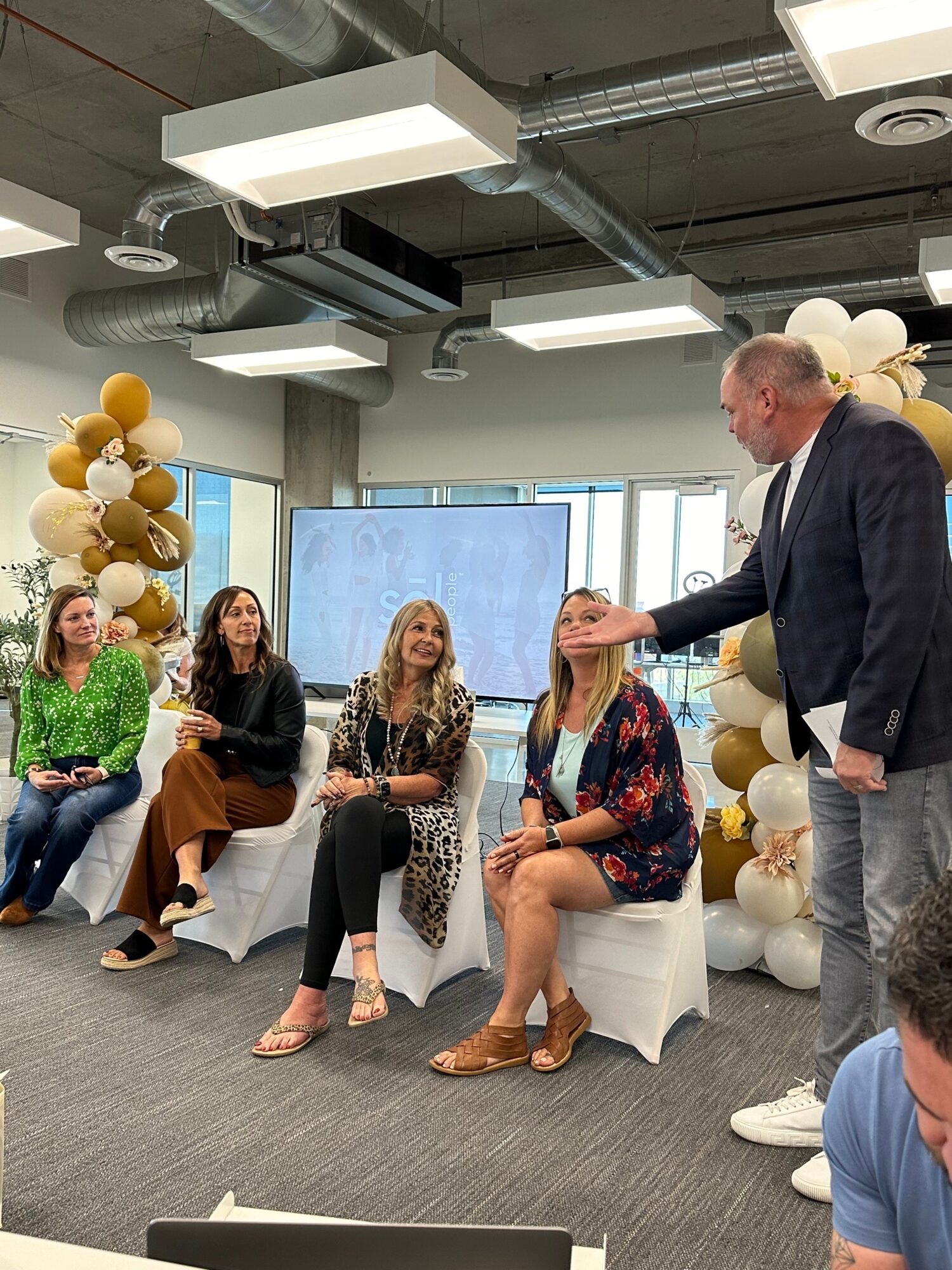 Man standing and speaking to four seated women in a modern room with balloon decorations and large windows.