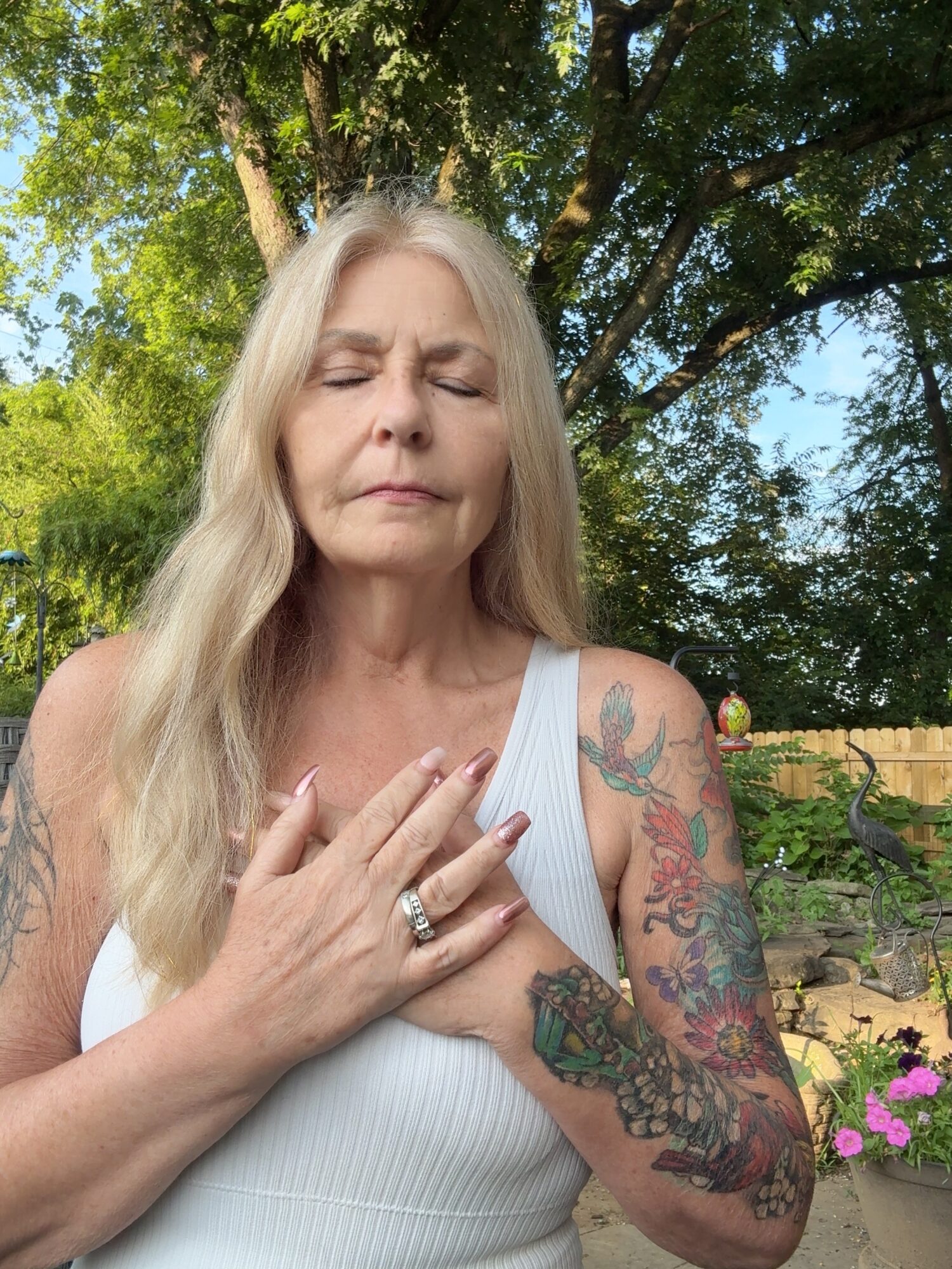 Woman with long blonde hair, eyes closed, touching chest, outdoors with trees and flowers.
