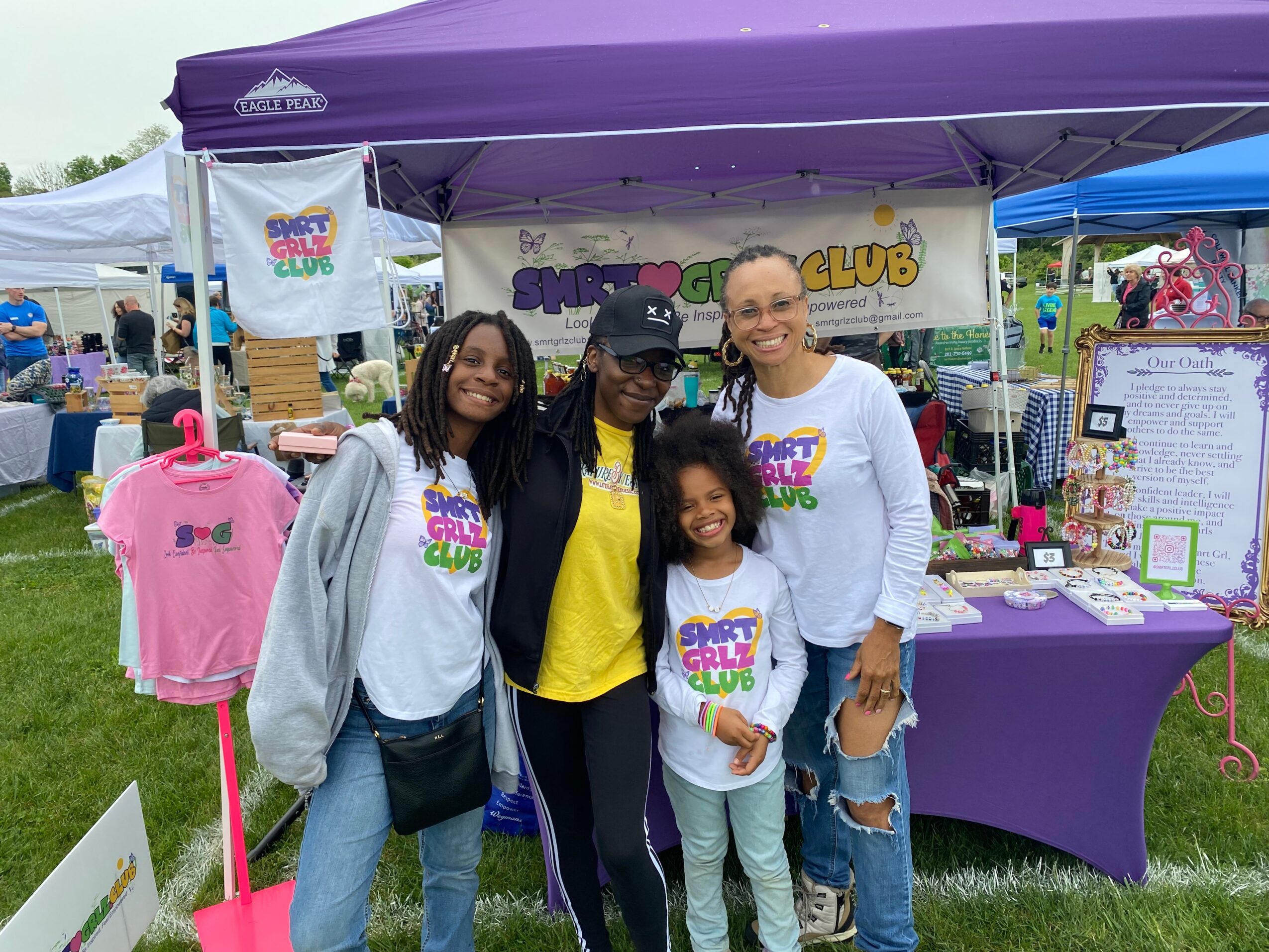 Four people smiling at an outdoor event, standing in front of a booth with purple tablecloth and banners.