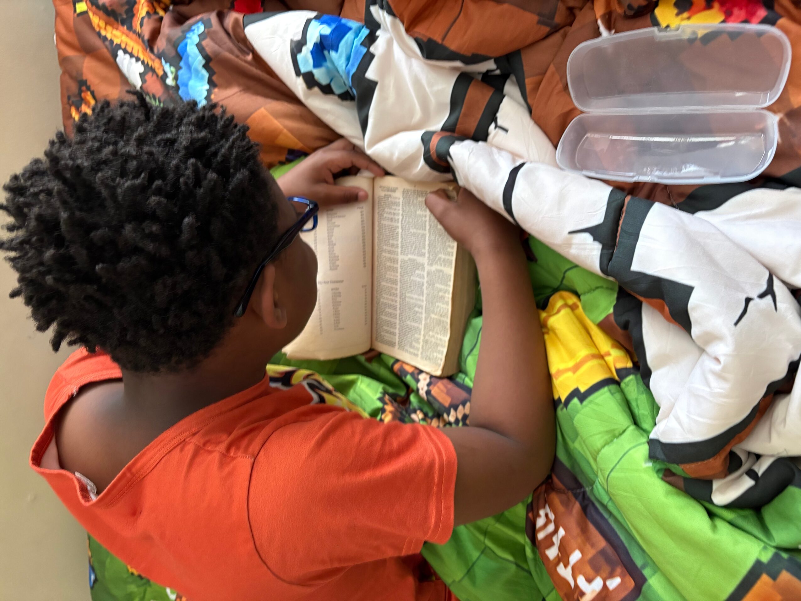 Child reading a book at a table with colorful items and a plastic container nearby.