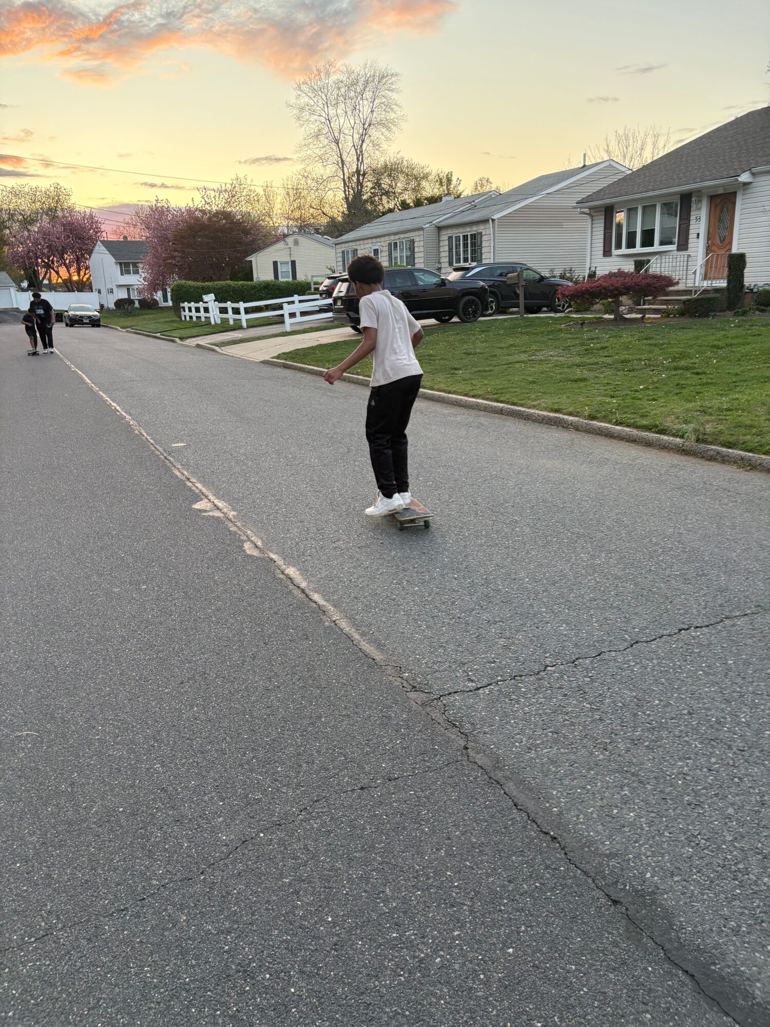 Teenager skateboarding on a suburban street with houses, cars, and trees in the background during sunset.