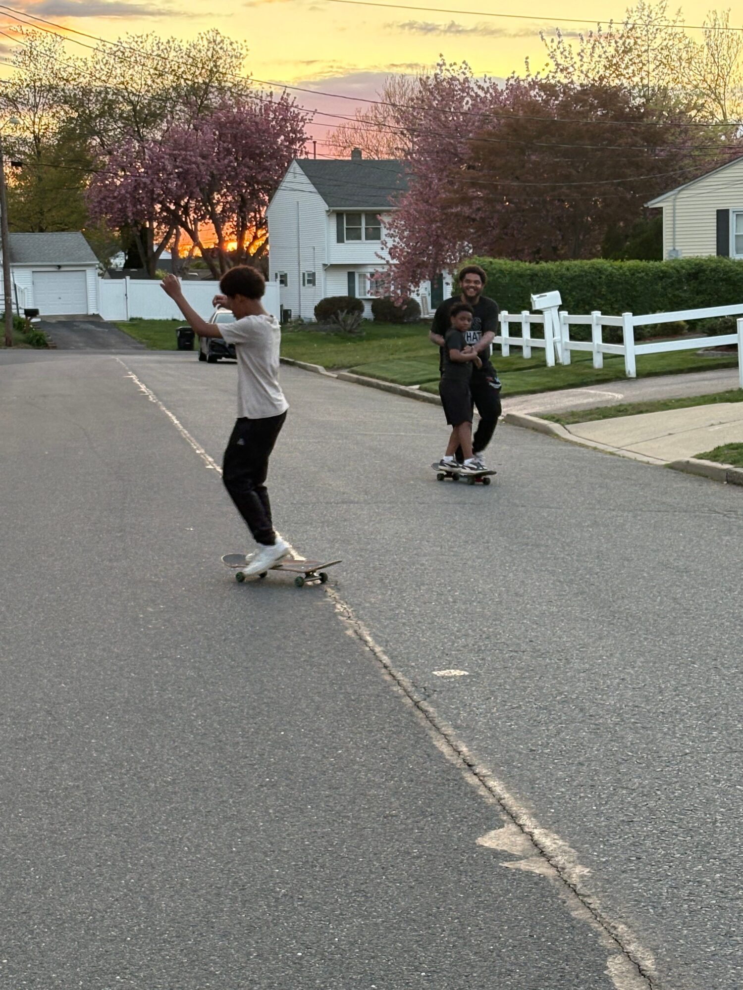 Two children skateboarding on a suburban street with houses and blooming trees in the background.