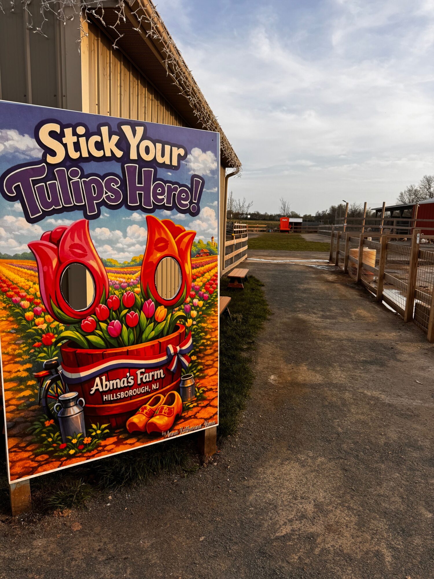 Sign with red sunglasses and flowers, advertising tulip viewing at Alma's Farm, with farm and sky in background.