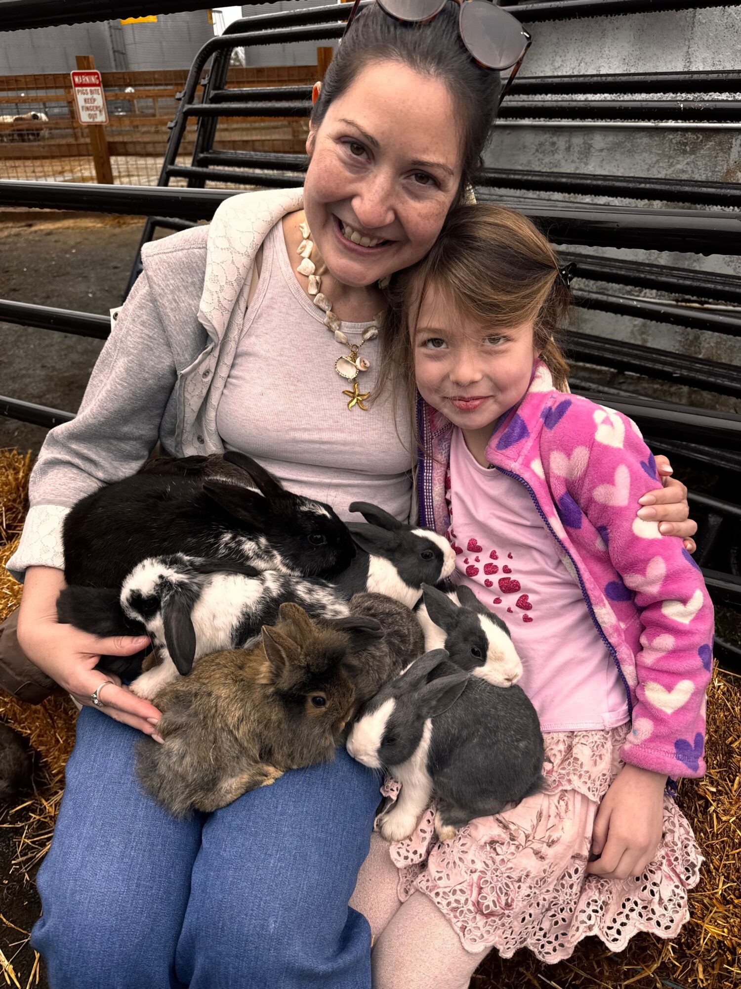 Woman and girl sitting with multiple rabbits outdoors, woman smiling, girl in pink jacket, black metal fence in background.