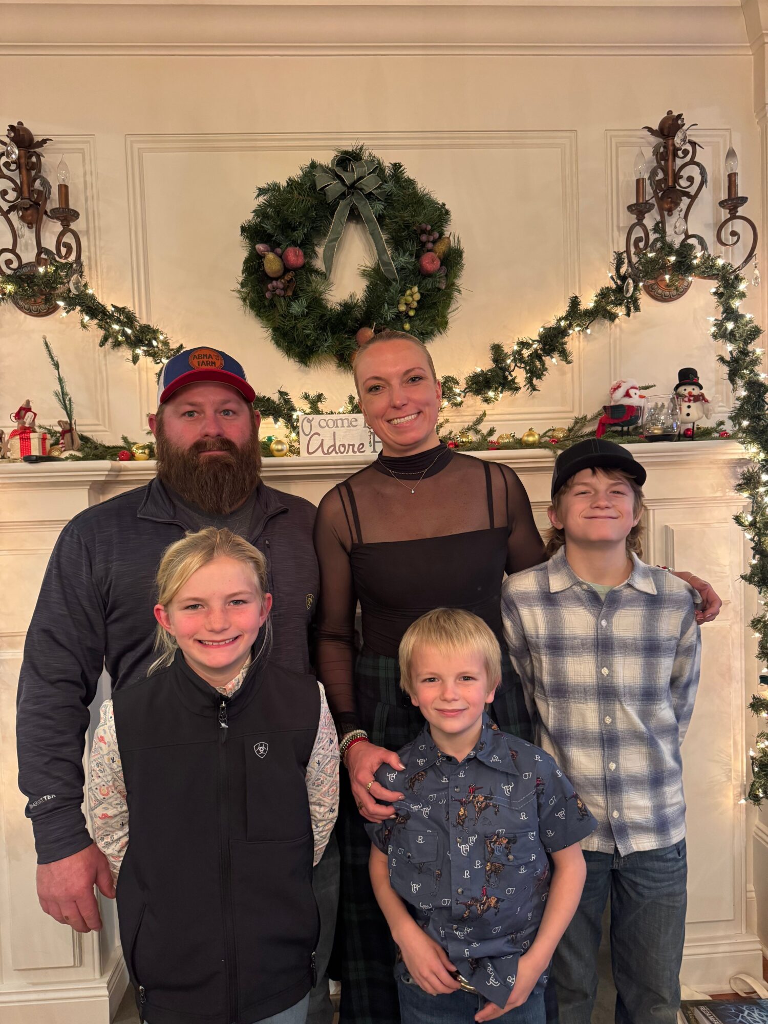 Group of five people, two adults and three children, standing in front of a decorated fireplace with a wreath and garland.