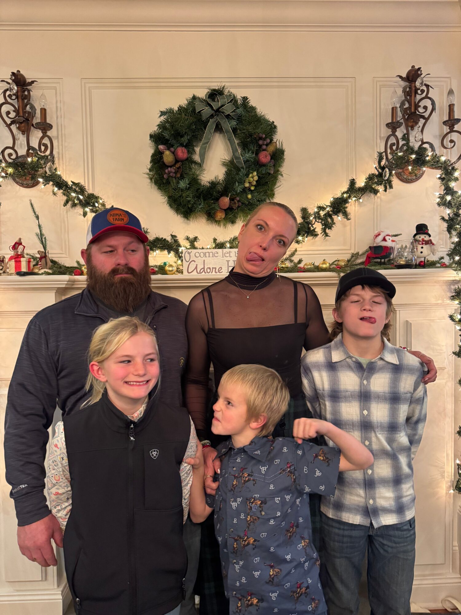 Group of five people, three children and two adults, standing in front of a decorated fireplace with a wreath and candles.