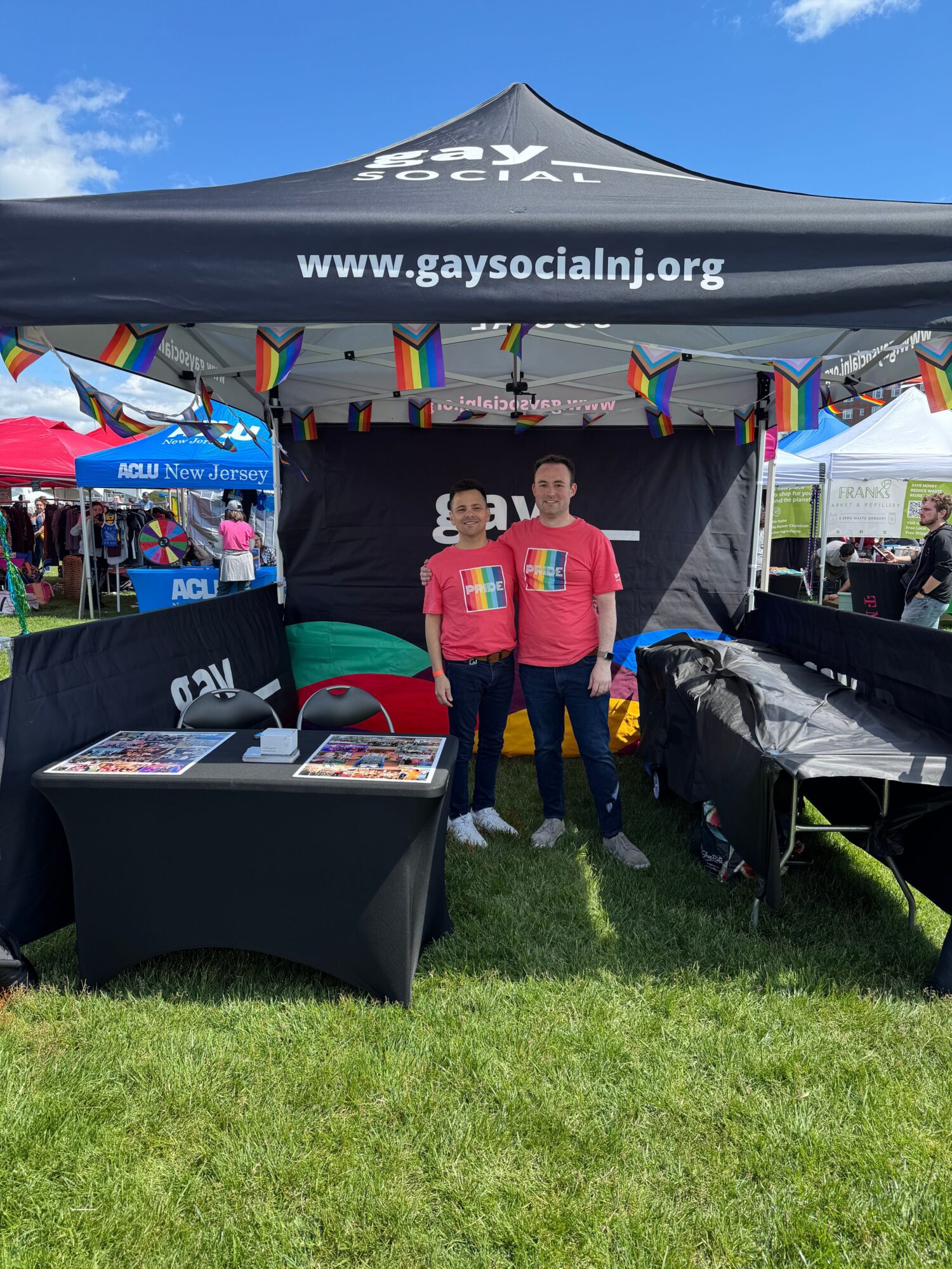 Two people standing under a black tent with rainbow flags, at an outdoor event with booths and tents.