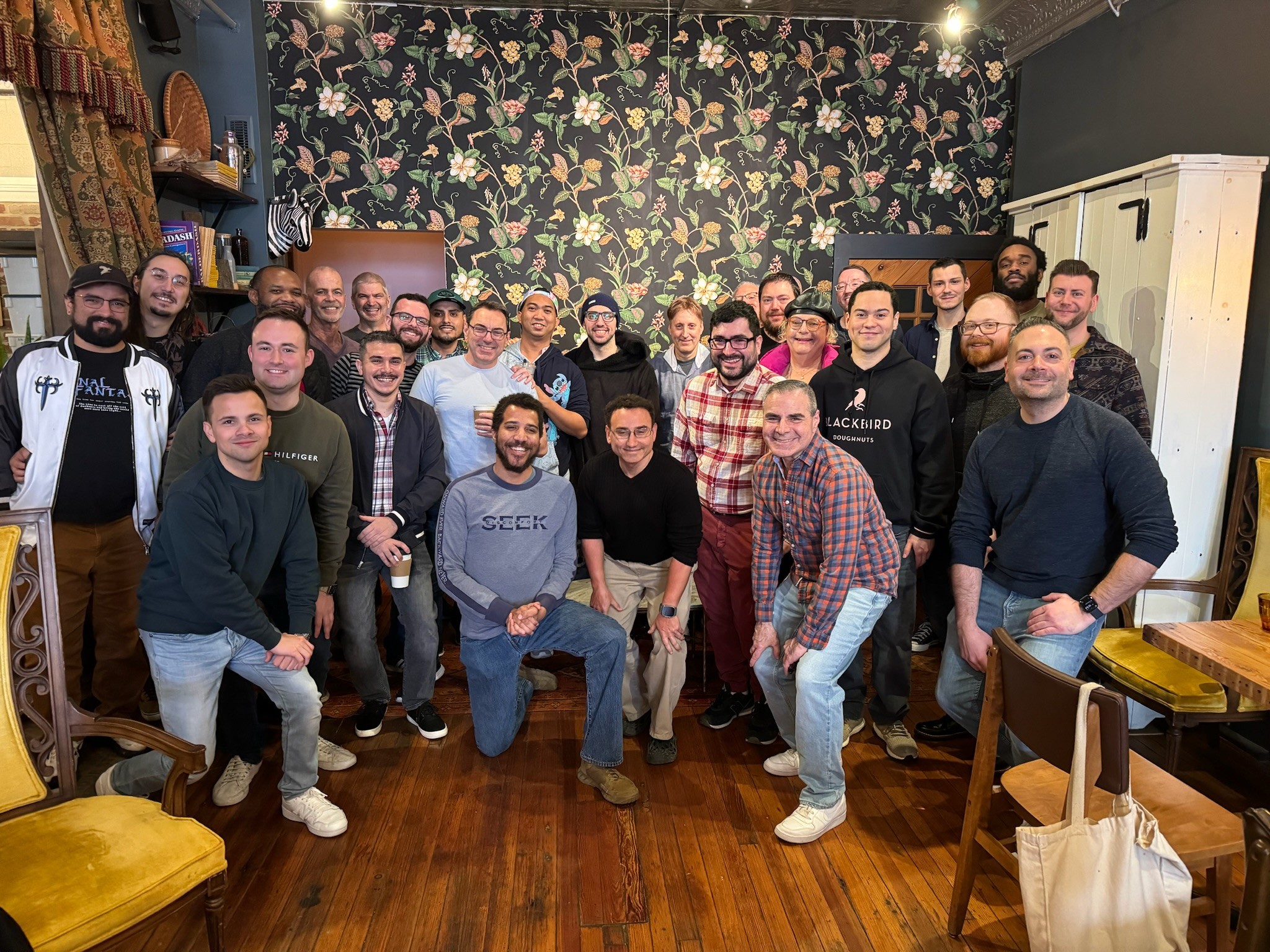 Group of 22 people gathered indoors, smiling, with a floral wallpaper background and wooden floor.