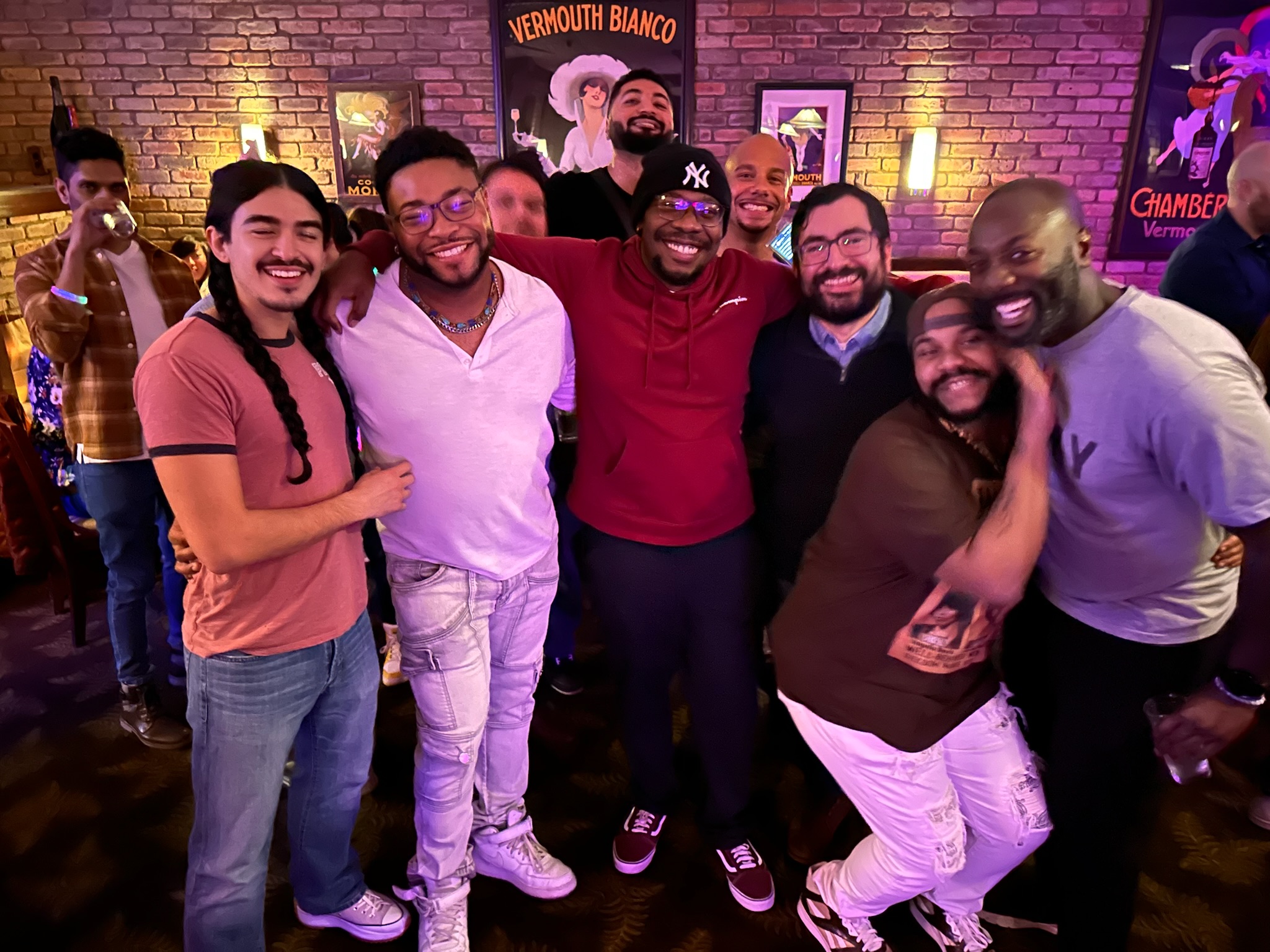Group of seven people smiling and posing together in a bar with brick walls and colorful signs.