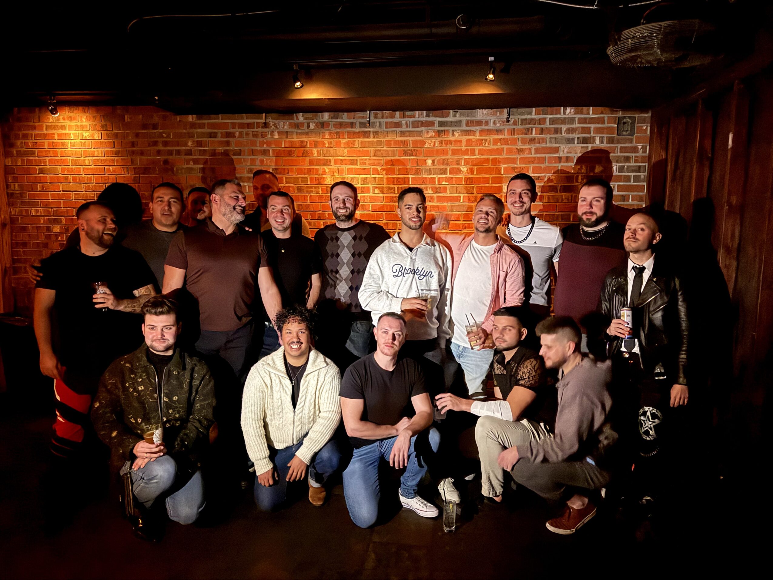 Group of 16 people posing in front of a brick wall, some holding drinks, in a dimly lit setting.