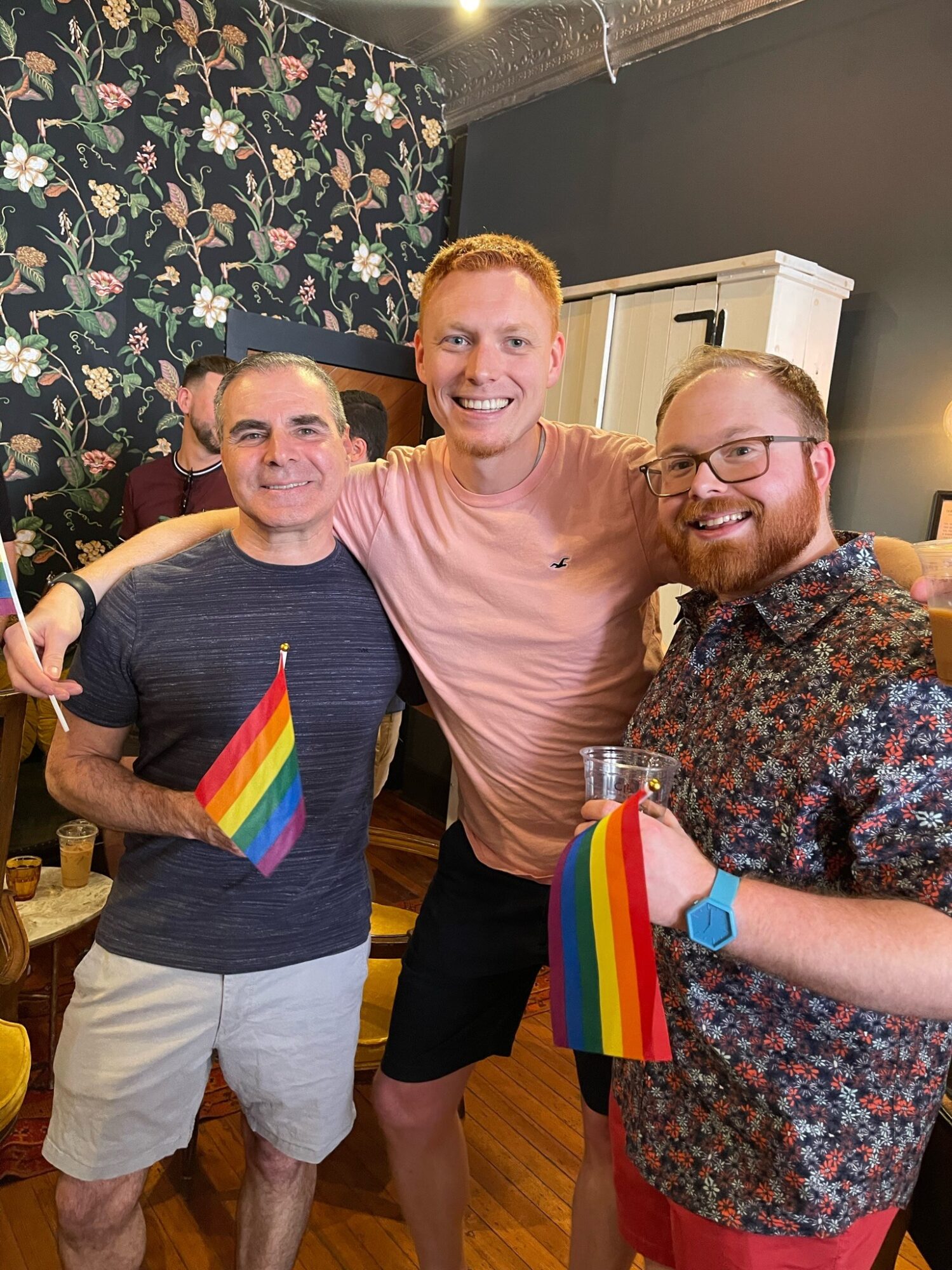 Three men smiling, holding rainbow flags, standing indoors with floral wallpaper and furniture in background.