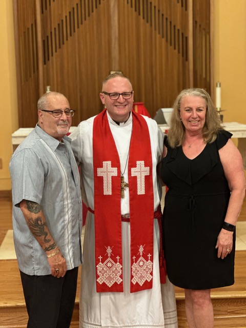 Three people standing together indoors, one in religious robes with a red stole, two in casual and formal attire.