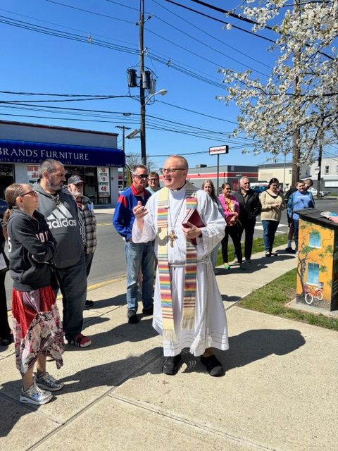 Clergy member in white robes and stole speaking to a group outdoors on sidewalk, with trees and power lines in background.