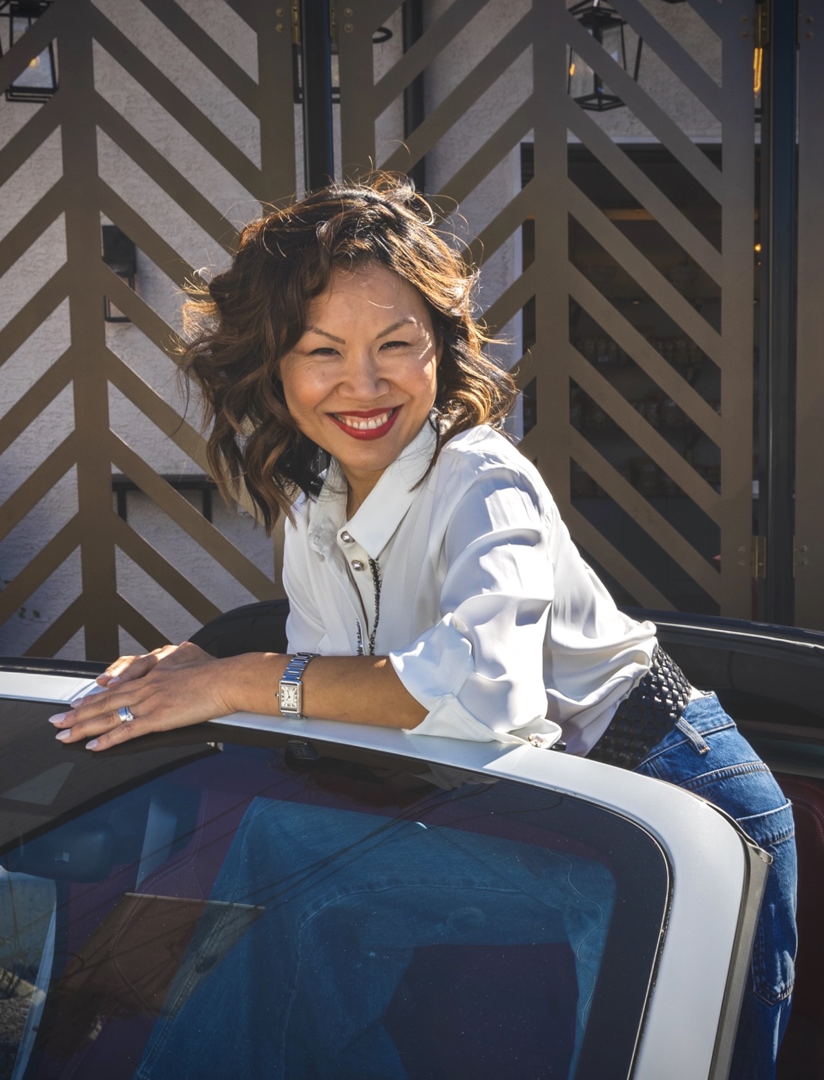 Woman with curly hair smiling, leaning on a car, outdoors with a wooden fence background.