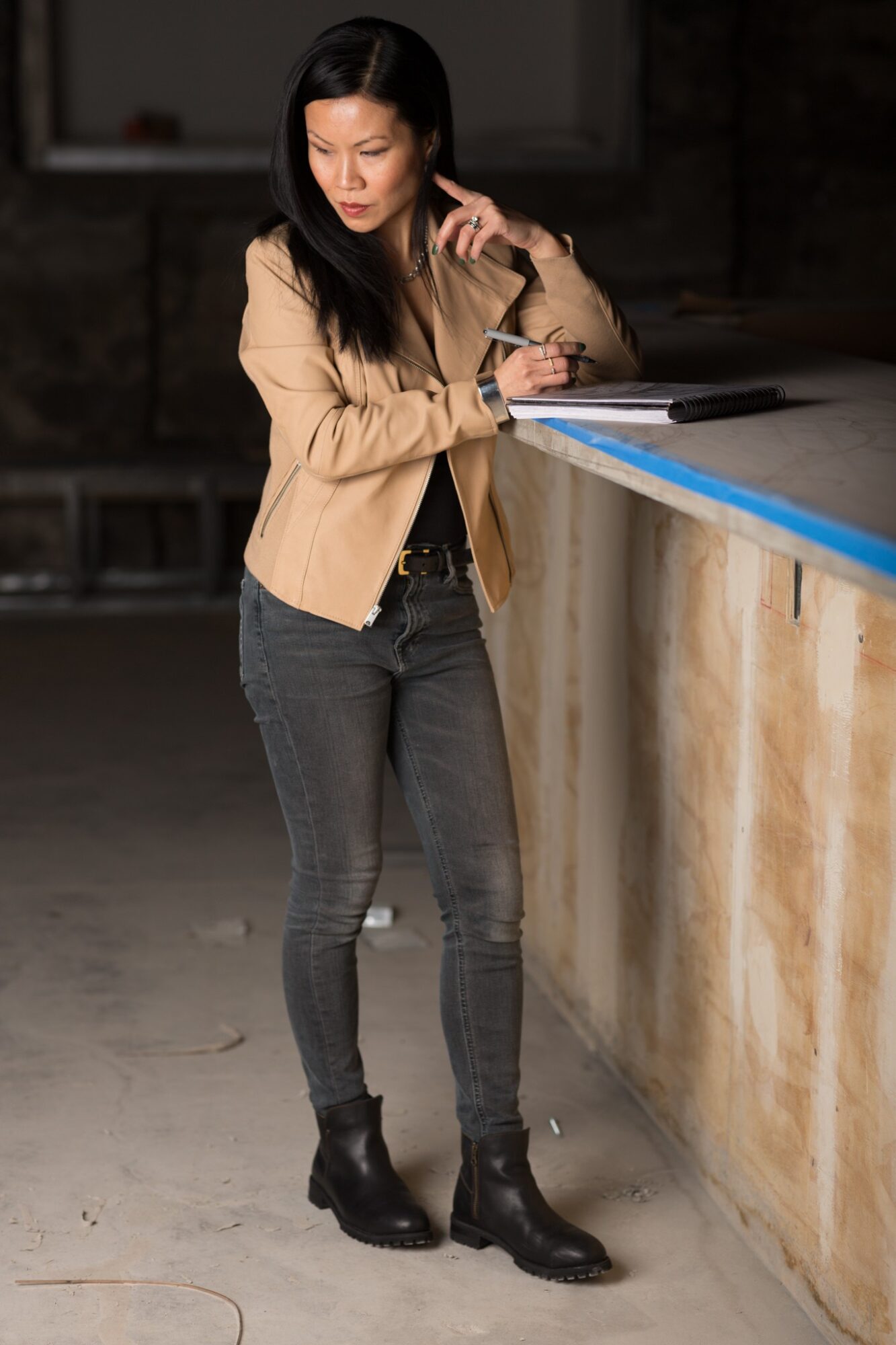 Woman standing at a counter, looking down, wearing a beige blazer, dark jeans, and black boots.
