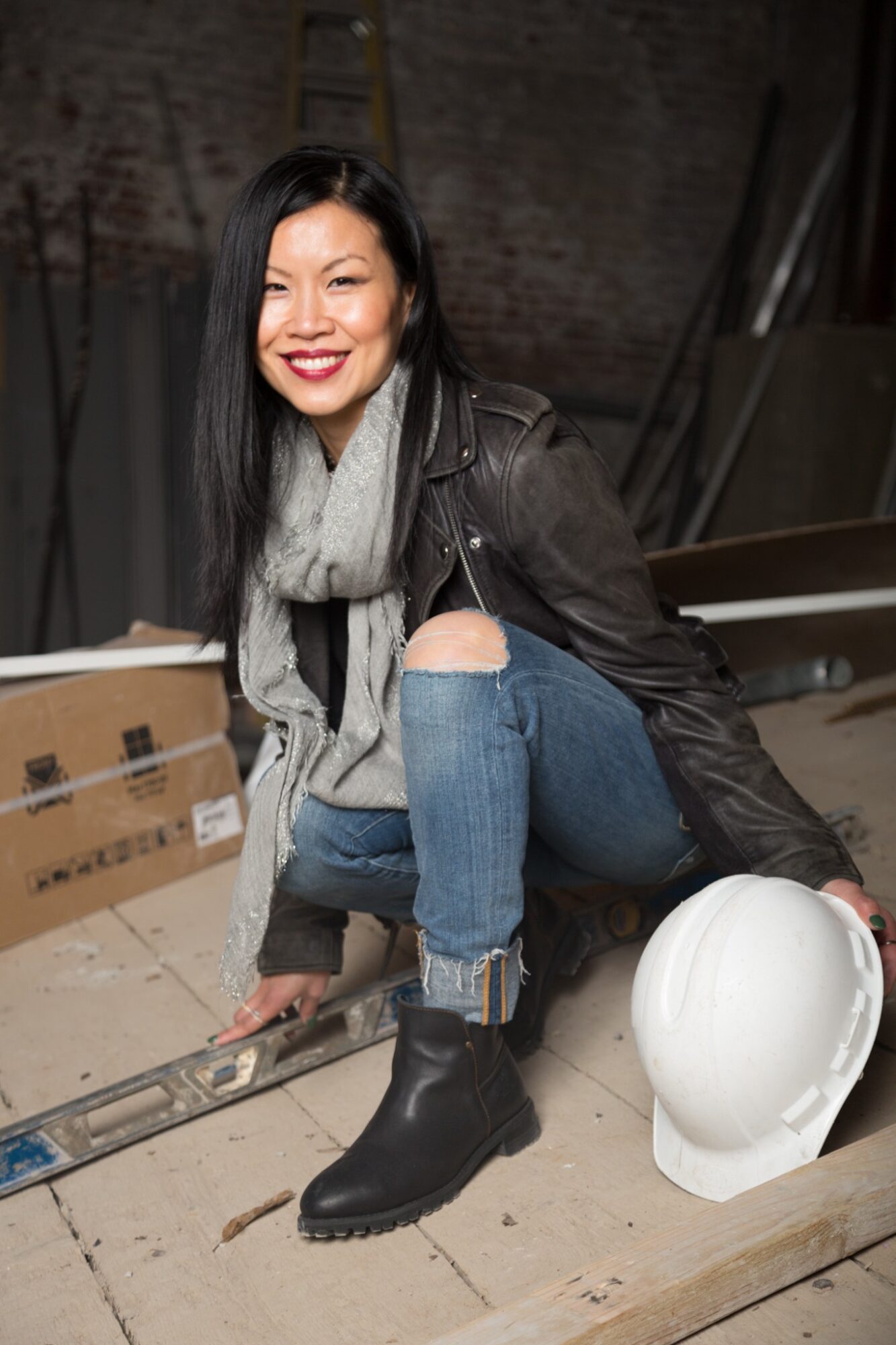 Woman with long dark hair smiling, wearing a leather jacket, gray scarf, ripped jeans, and black boots, sitting on floor with a white helmet nearby.