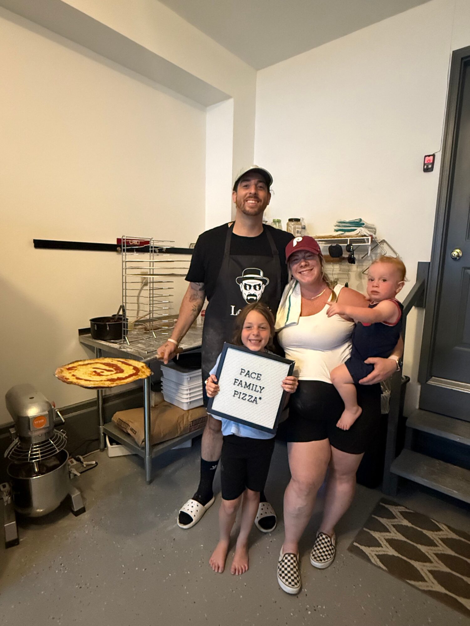 Group of four people, two adults and two children, standing indoors, smiling, with a sign reading 'PEACE FAMILIES PRAY'.
