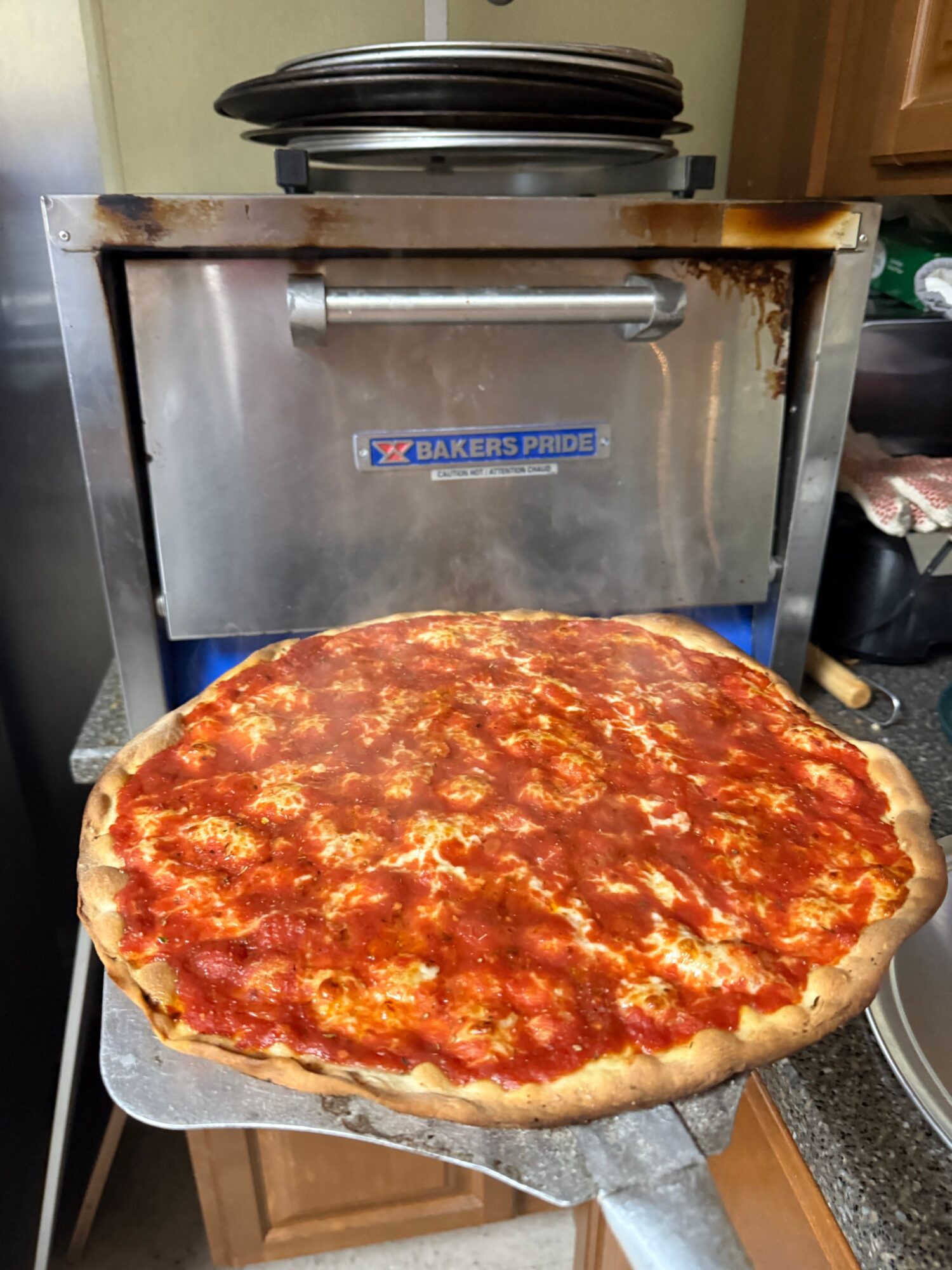Uncooked pizza with tomato sauce on a wooden countertop, in front of a stainless steel oven, with stacked black pans on top.
