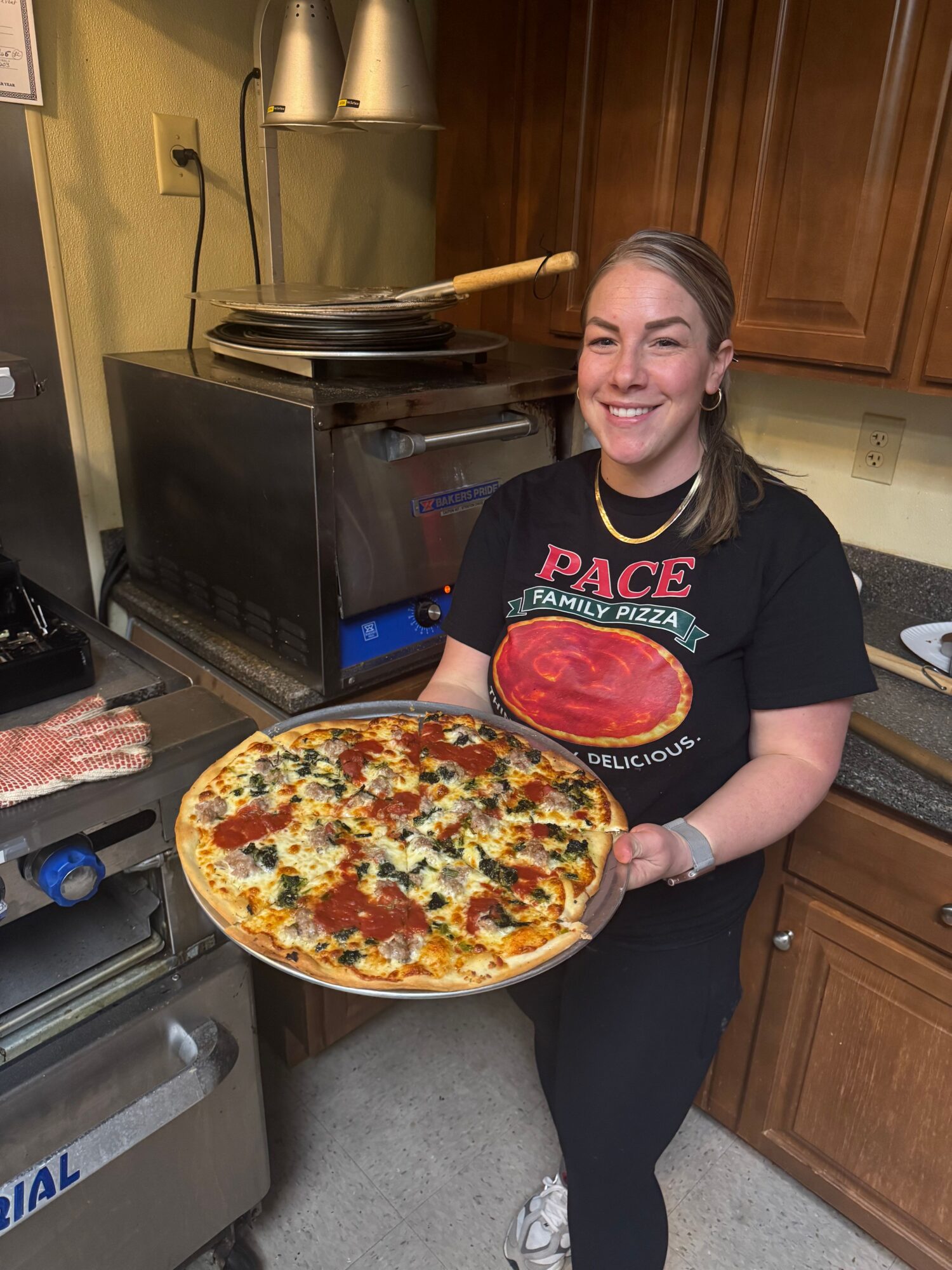 Smiling woman holding a large pizza in a kitchen with wooden cabinets and cooking equipment.