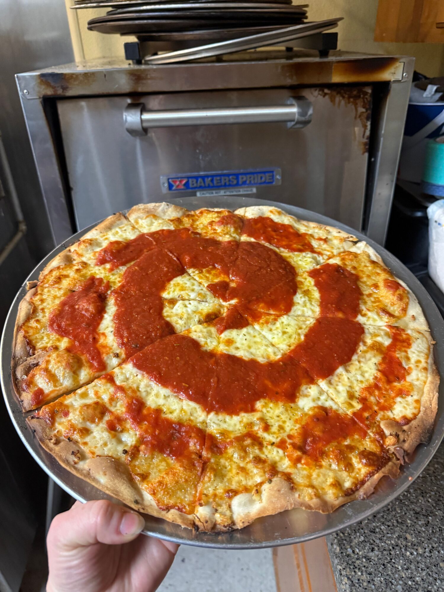 Pizza with cheese and tomato sauce on a metal tray, held by a hand.