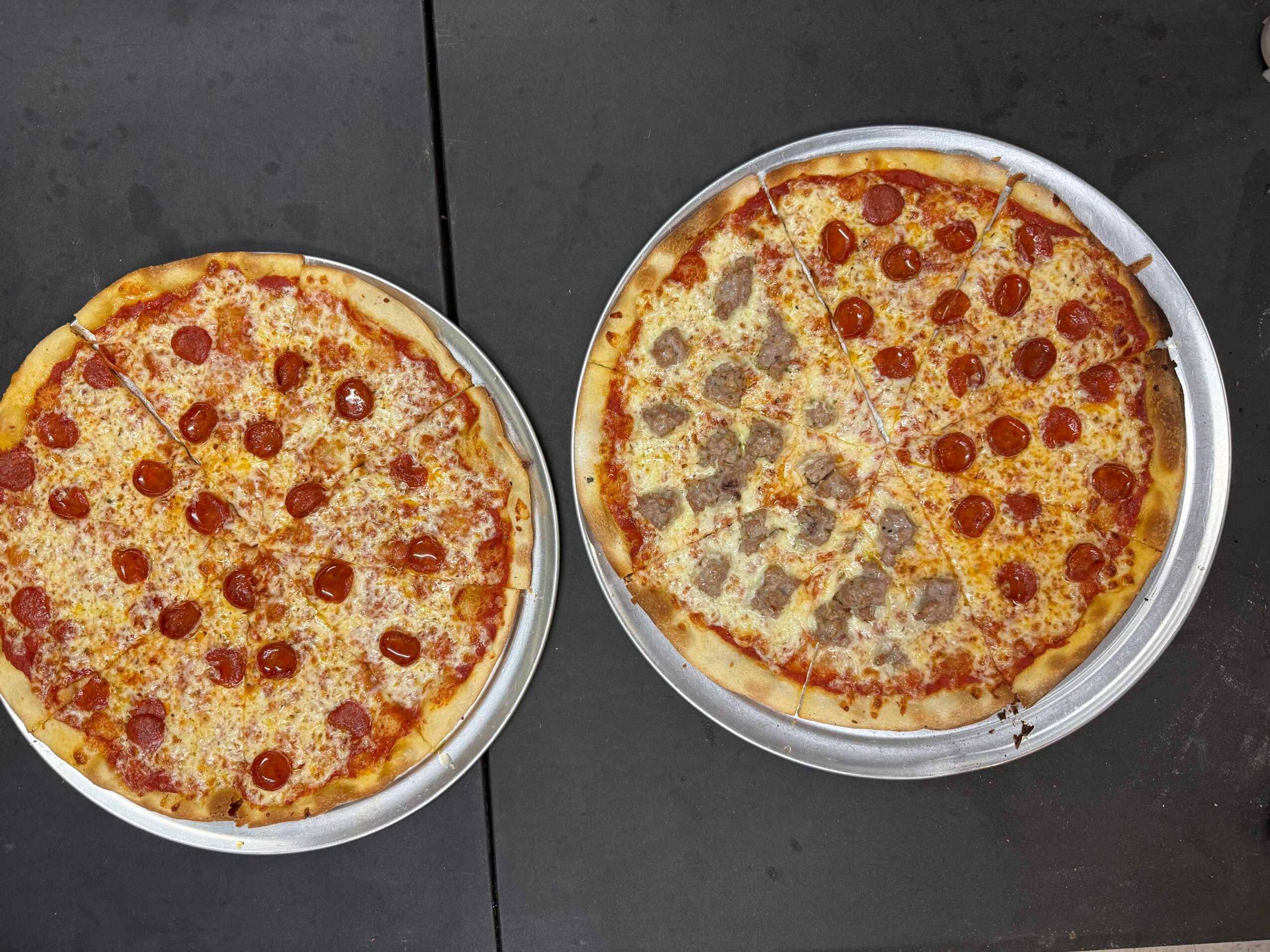 Two pizzas on metal trays, one with pepperoni and cheese, the other with cheese and toppings, on a dark surface.