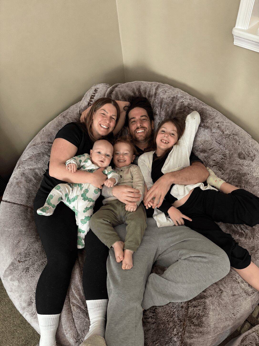 Four people, two adults and two children, lying on a gray couch, smiling and hugging each other.